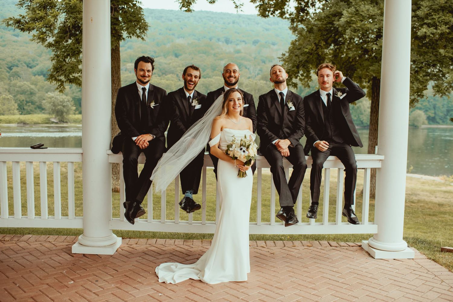 Wedding party poses on elegant white columned porch overlooking scenic mountain landscape.