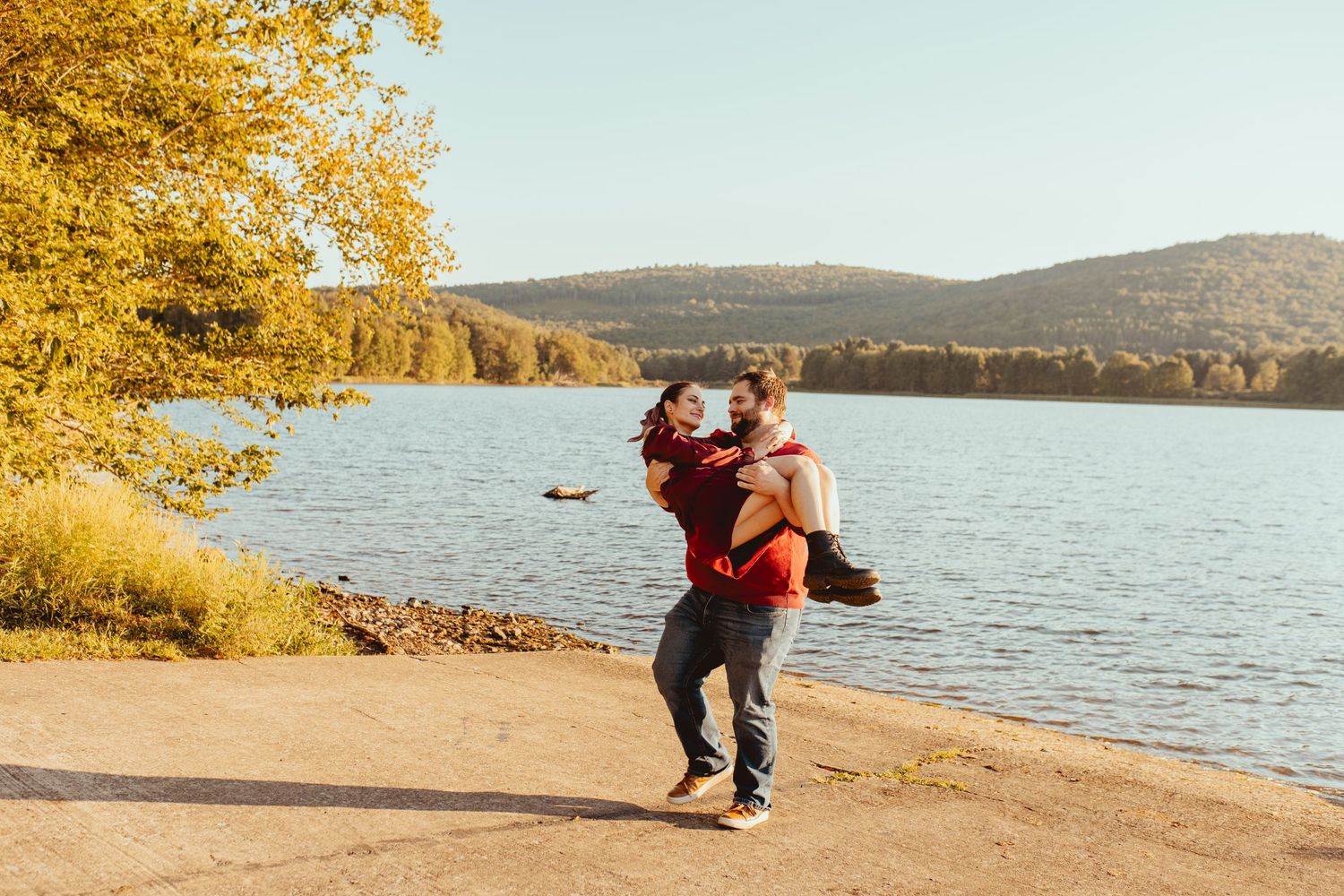 A person in a red jacket and black pants walks along a scenic autumn lakeside trail during golden hour.