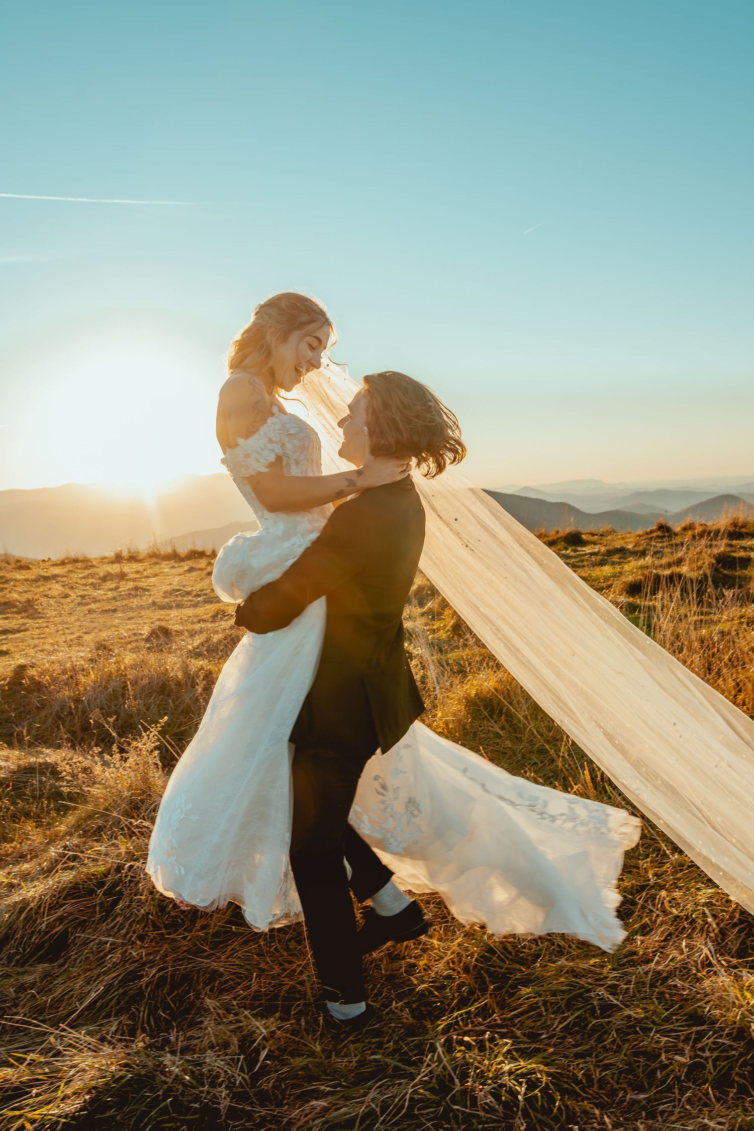 A romantic wedding couple embraces on a hilltop at sunset with a flowing veil catching the golden light.