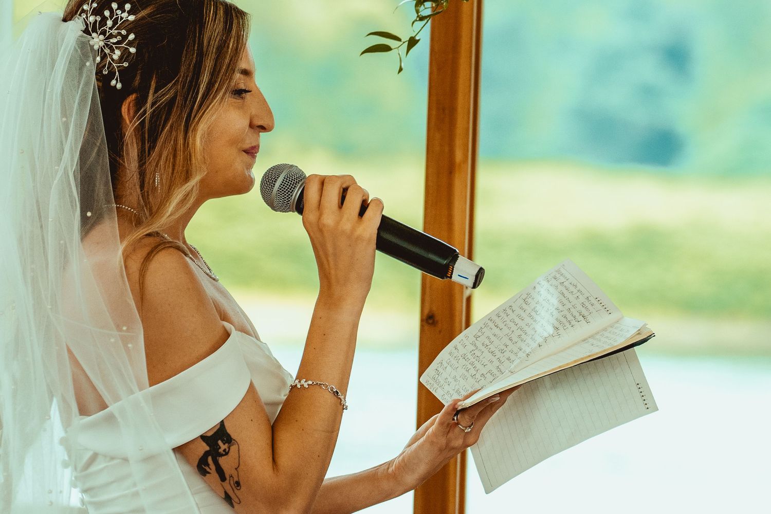 Bride in a white dress gives a heartfelt speech while holding a microphone and paper at an indoor wedding venue.