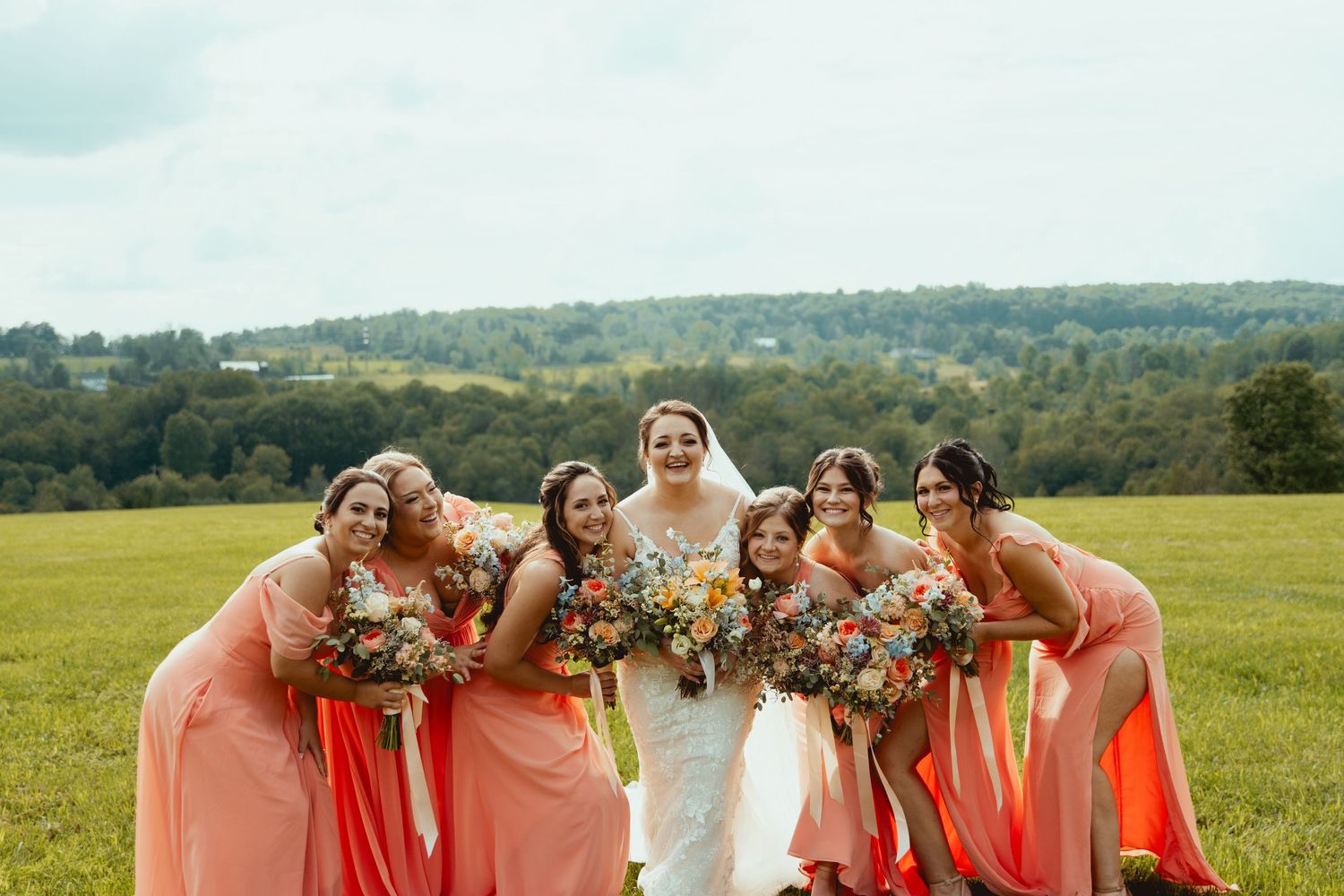 Bride in white gown with six bridesmaids in coral dresses holding colorful bouquets in a green field.