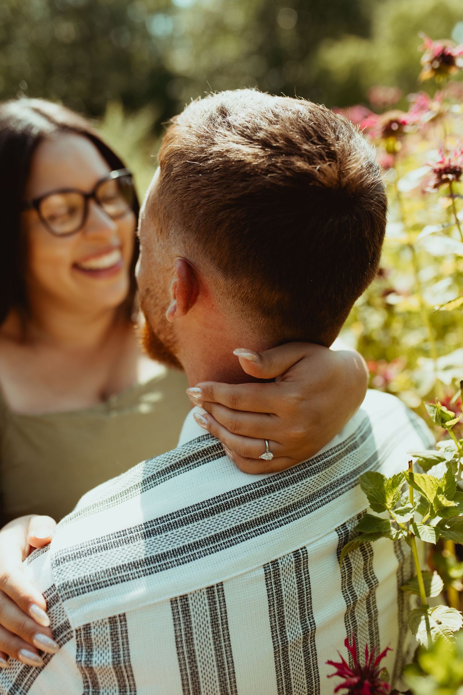 Smiling woman with glasses embraces man outdoors among red flowers, showcasing engagement ring on his neck.
