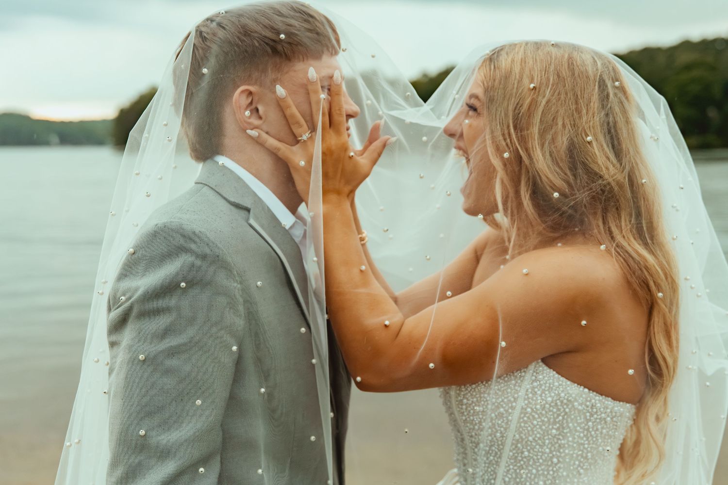 Romantic couple shares intimate moment under pearl-adorned wedding veil by lakeside at sunset.