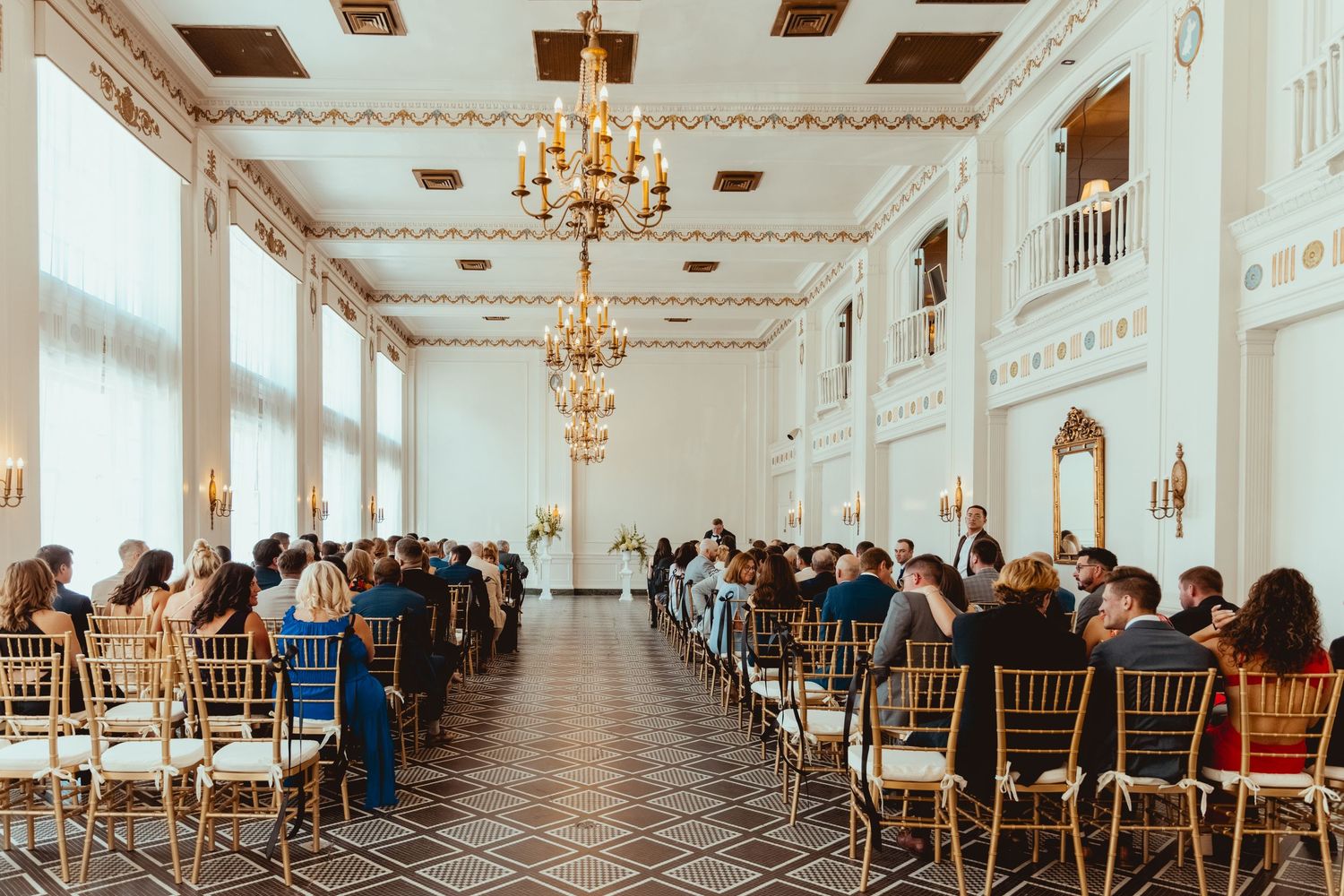 Elegant wedding ceremony in a grand white ballroom with ornate chandeliers and rows of wooden chairs.