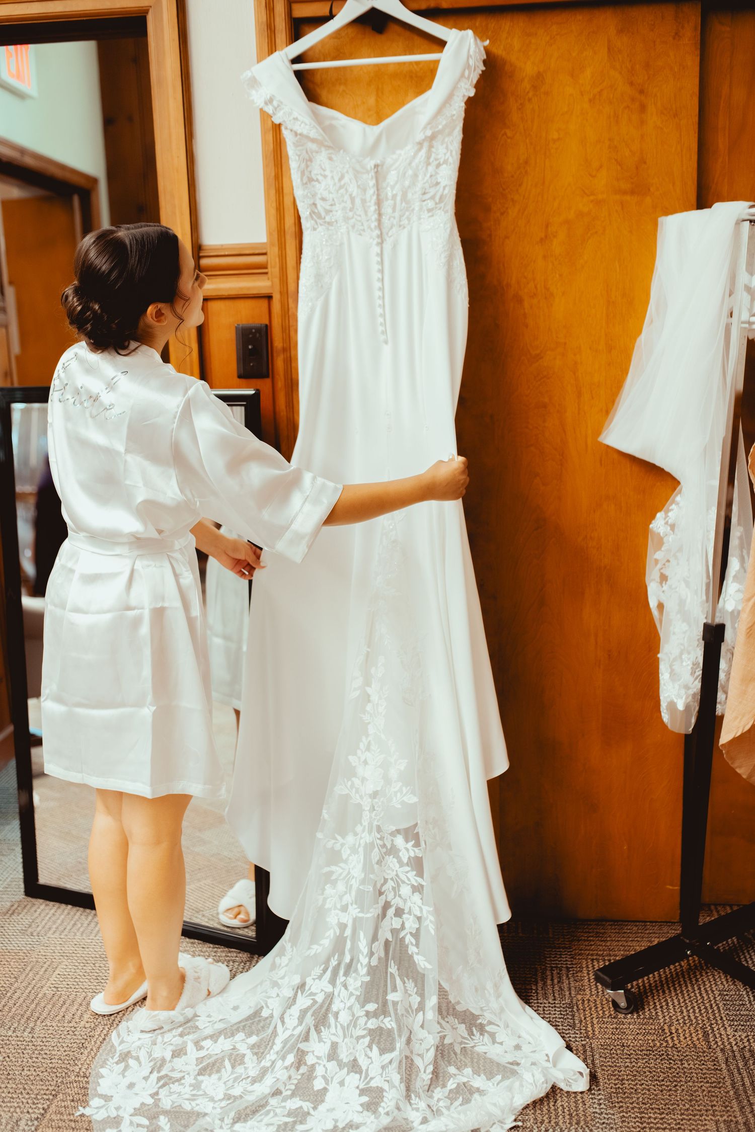 A beautiful white lace wedding dress with a long train hangs on display as someone in a white robe admires it.