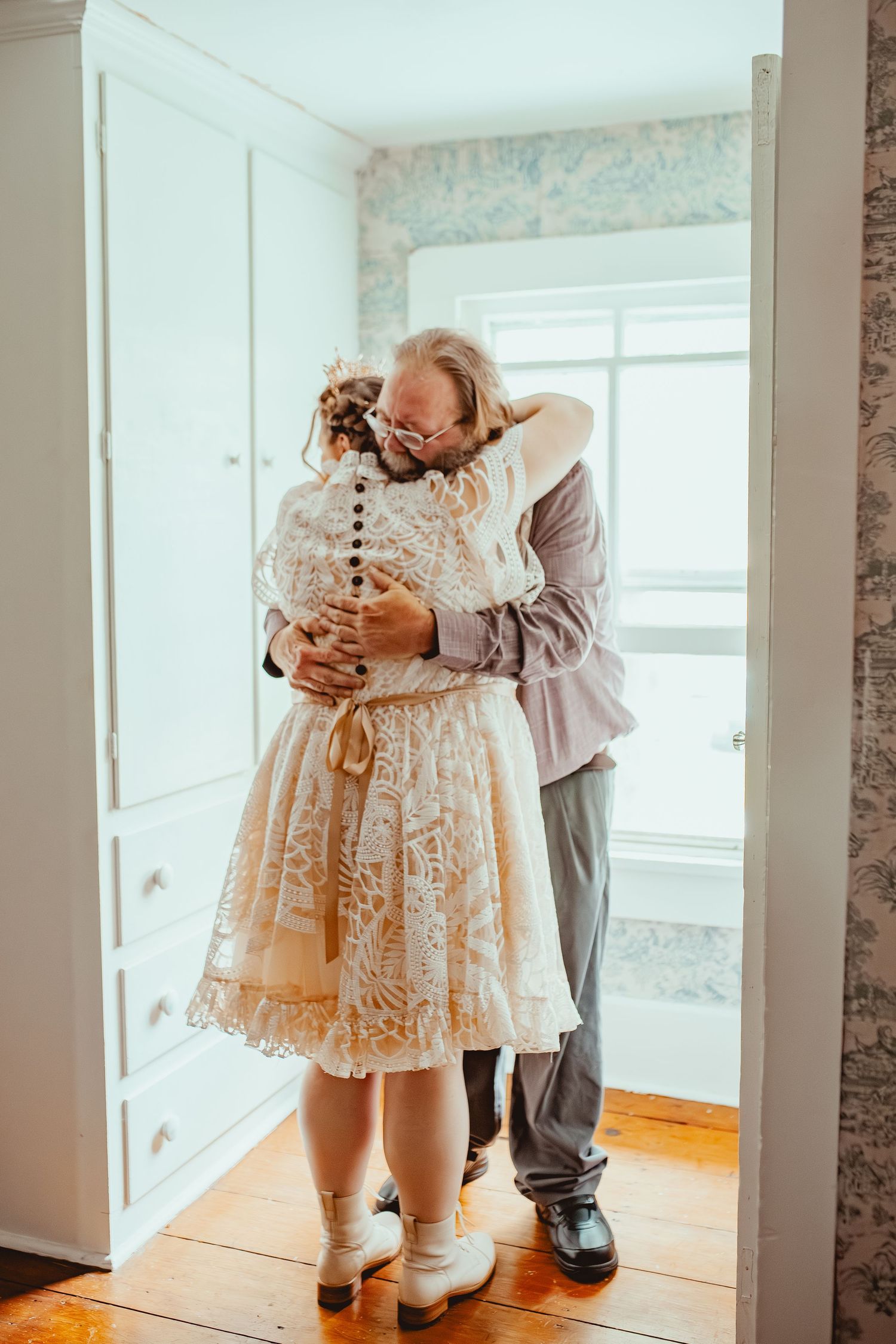 Two people share an intimate embrace in a room with floral wallpaper and hardwood floors.