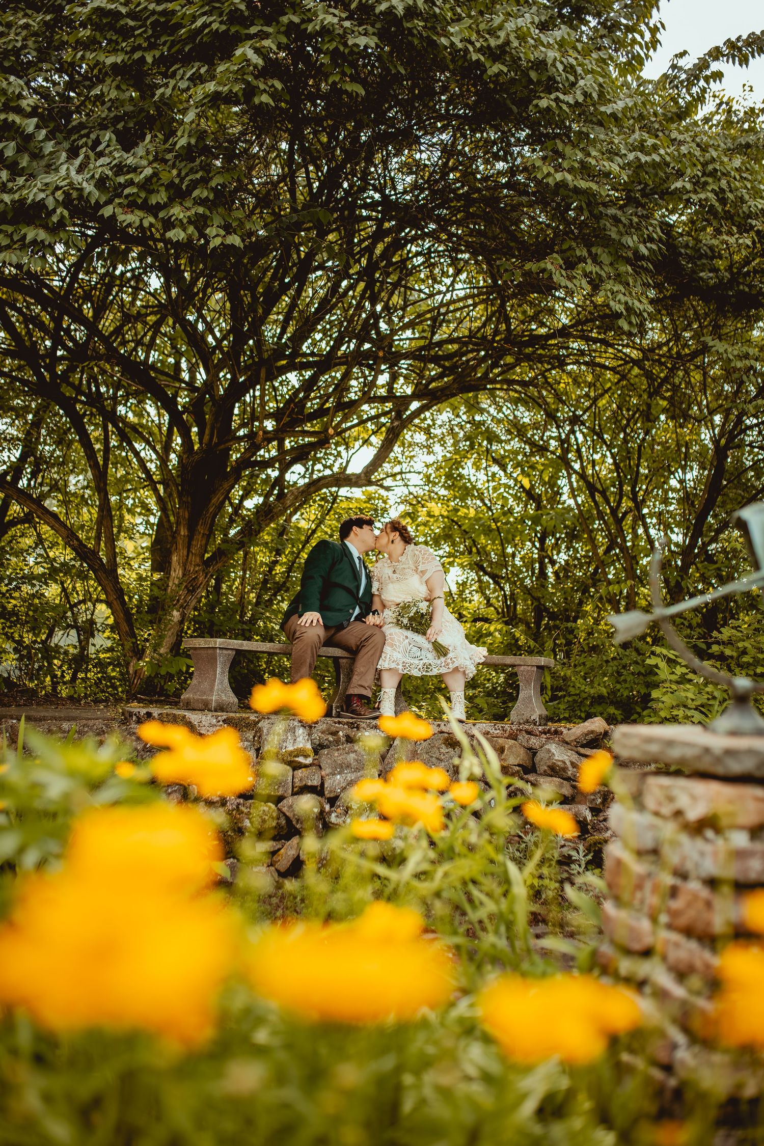 A couple sits on stone wall surrounded by yellow flowers and trees in a romantic garden setting.