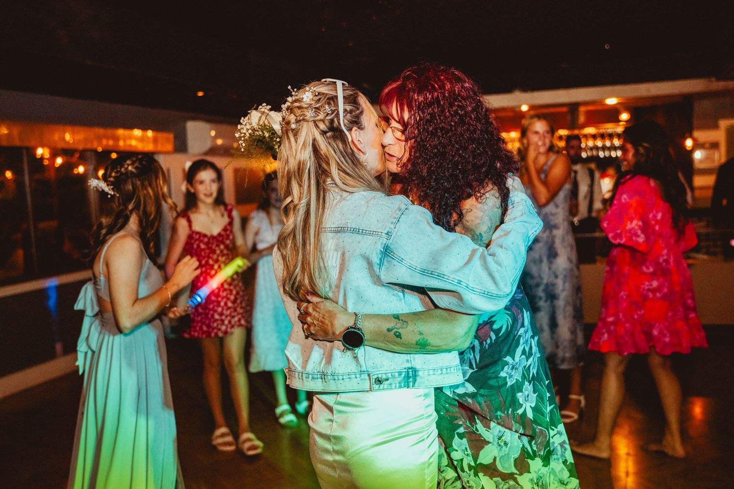 People dancing and celebrating at a dimly lit wedding reception with colorful lighting and festive atmosphere.