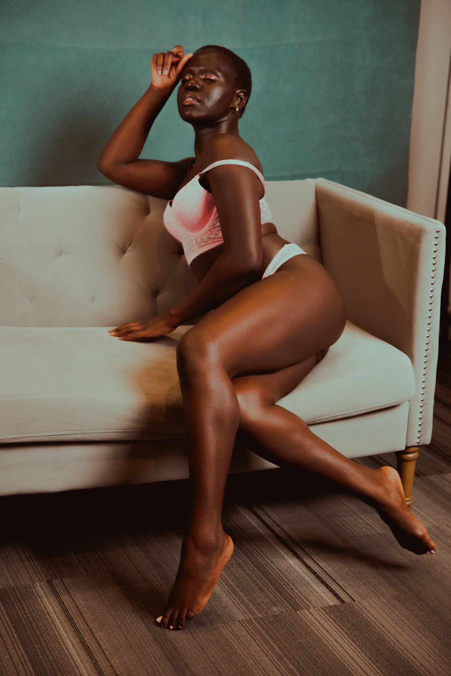 A woman in white lingerie poses gracefully on a cream tufted sofa against a teal wall.