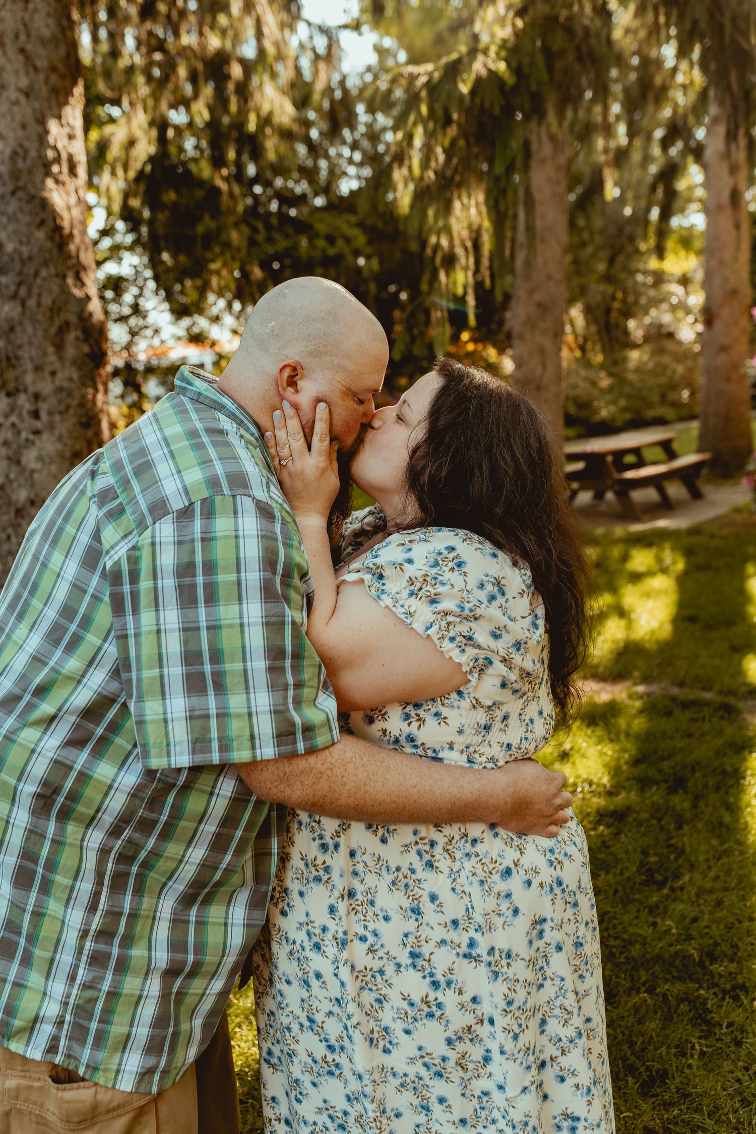 A couple shares a tender kiss outdoors among trees, the man in plaid and woman in a floral dress embracing warmly.