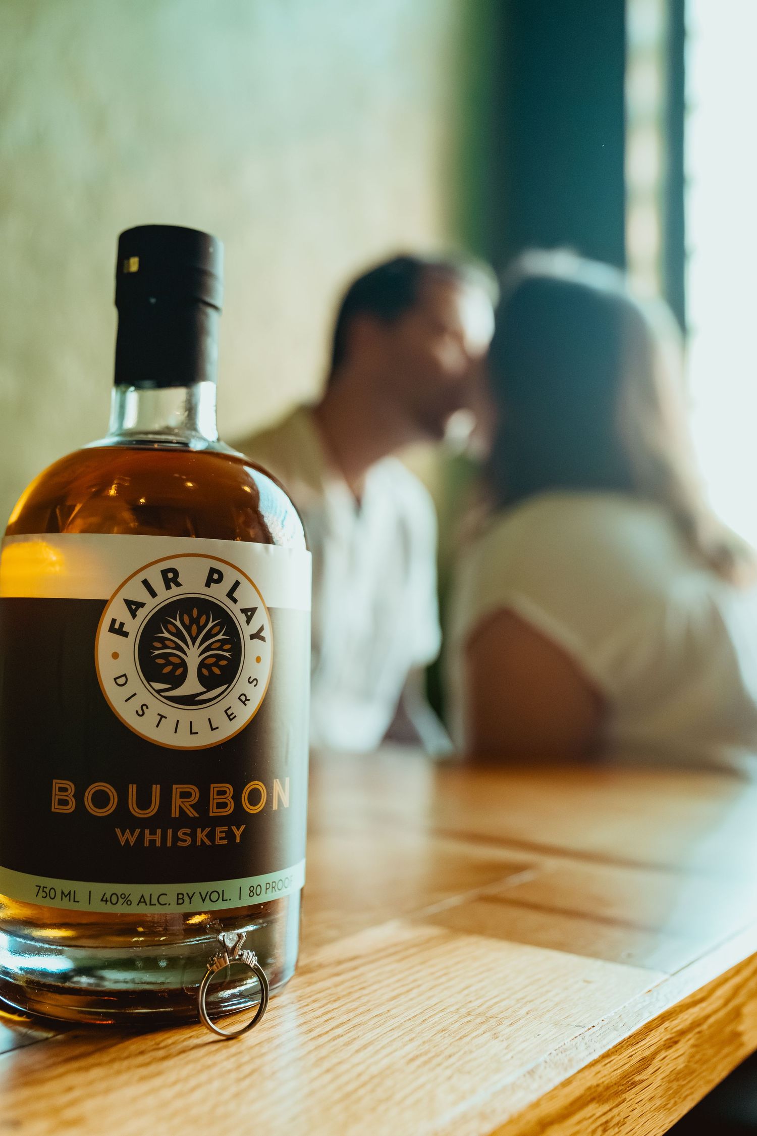 A bottle of Fair Play Distillers Bourbon Whiskey on a table with an engagement ring and a couple kissing in the background.