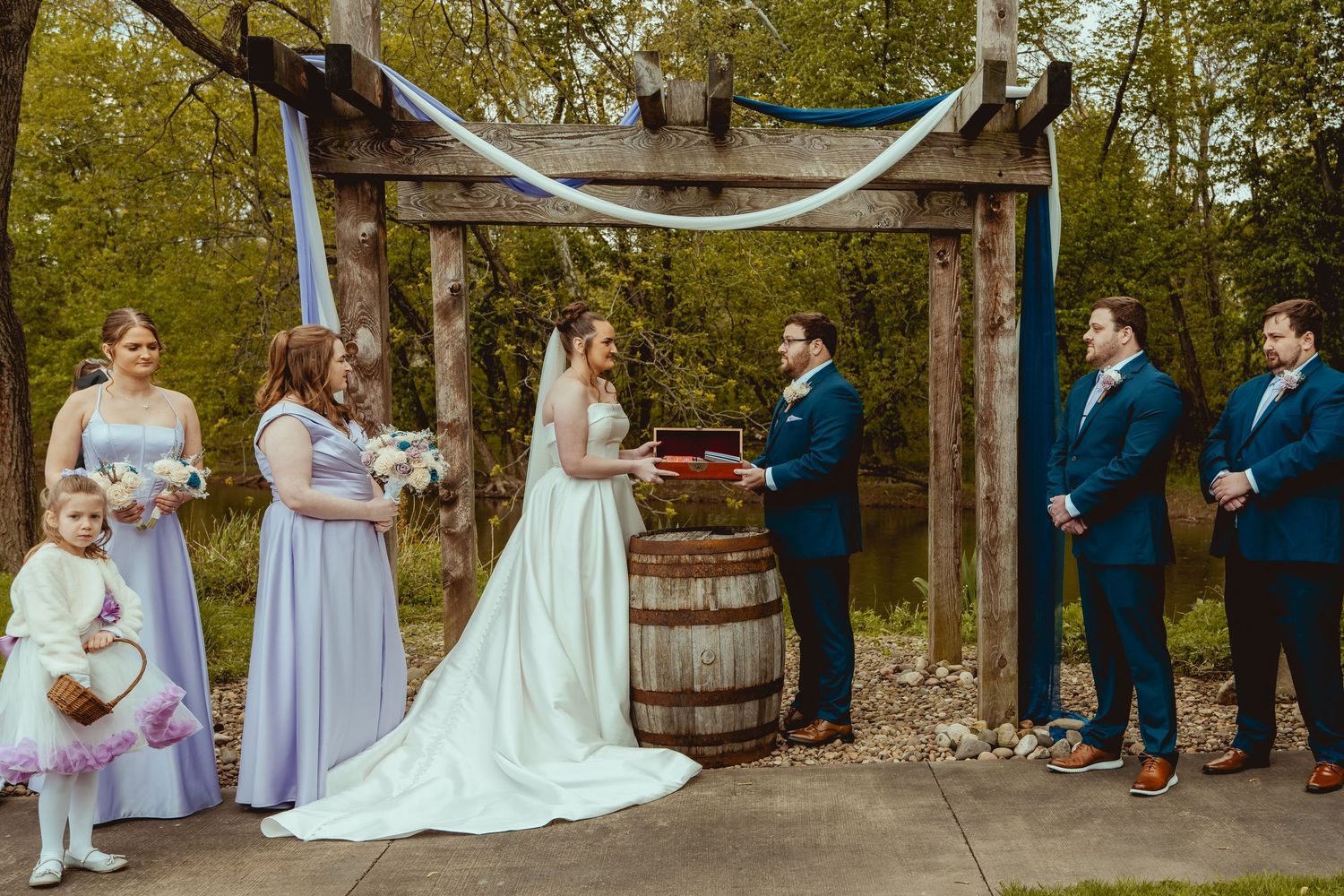 Outdoor wedding ceremony under rustic wooden arch with bridal party beside a pond.
