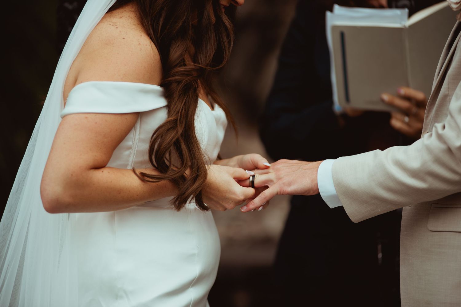 Bride and groom exchanging rings during wedding ceremony, bride in off-shoulder white gown with flowing veil.