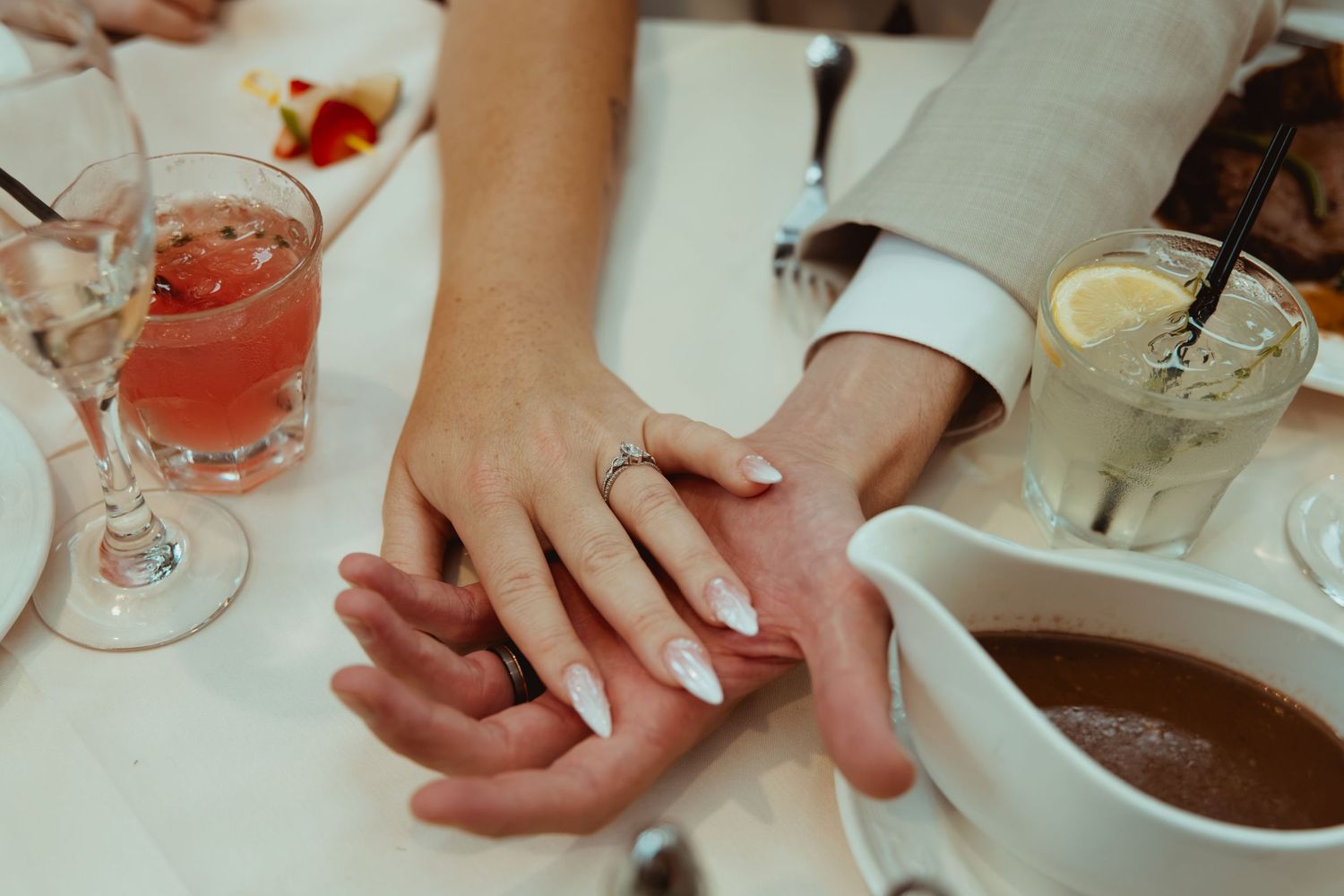 Couple holds hands displaying wedding ring on elegant table setting with colorful cocktails and gravy boat.