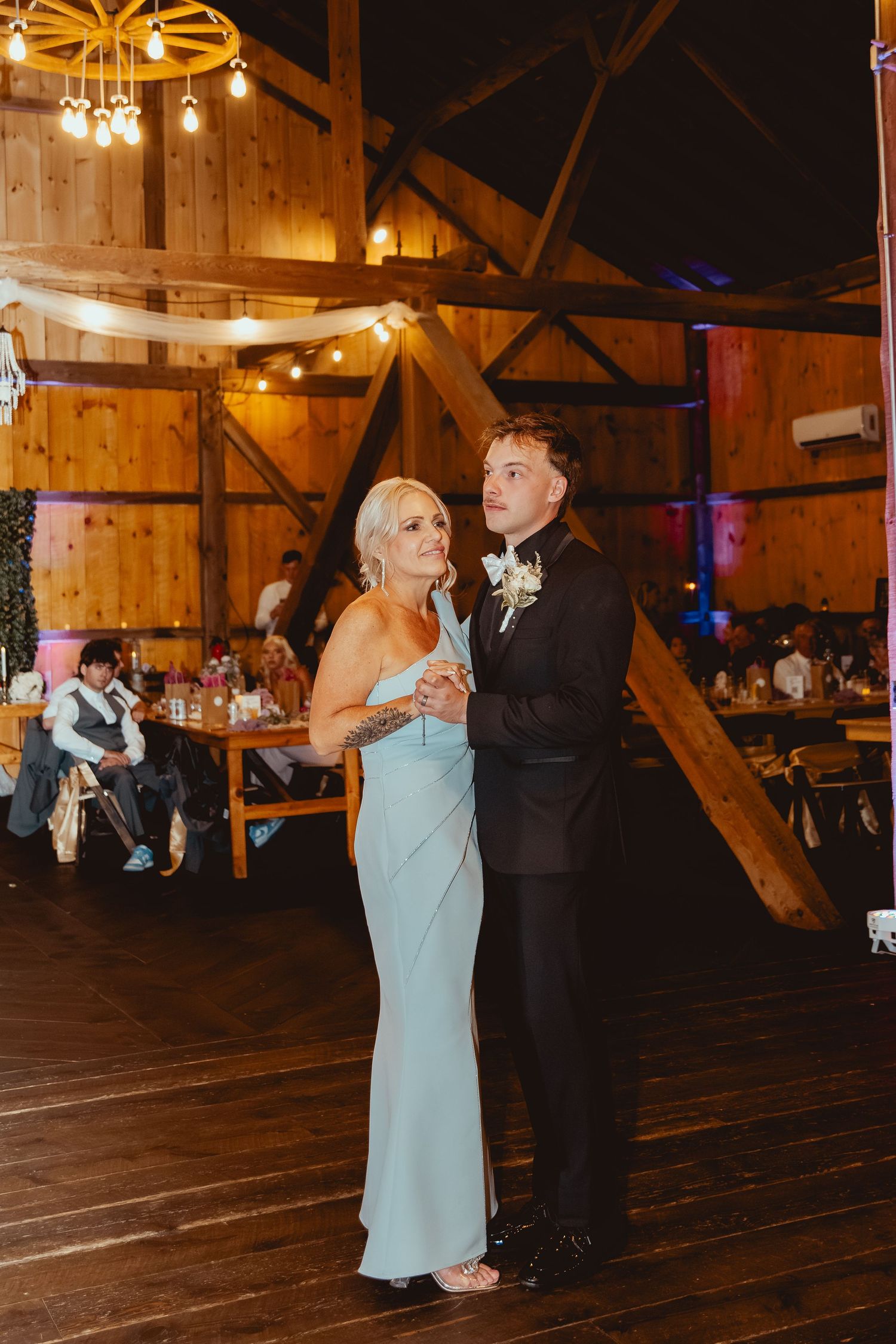 Wedding reception dance in rustic barn venue with hanging lights and wooden beams.