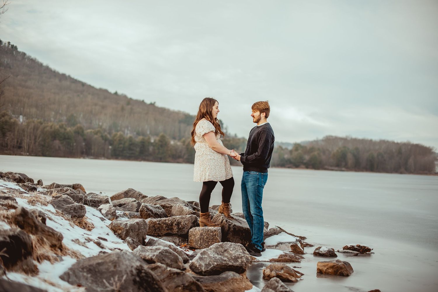 A couple stands together on rocky shoreline overlooking a misty lake and mountains.
