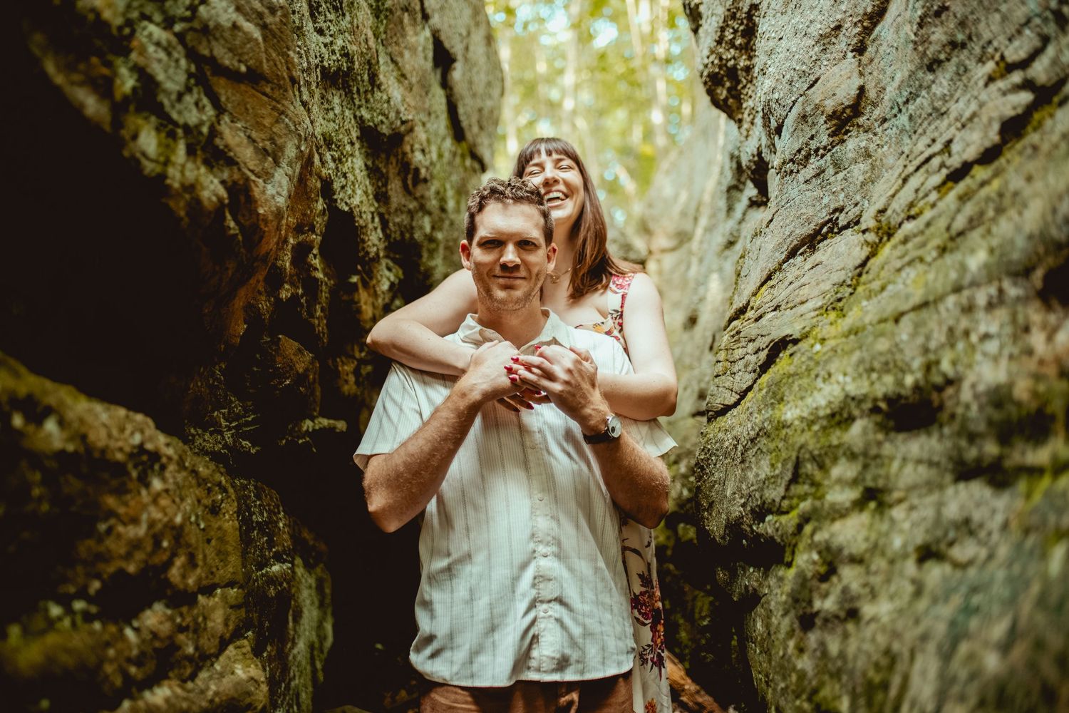 Couple embracing between mossy rock formations in a forest, woman hugging man from behind, smiling joyfully.