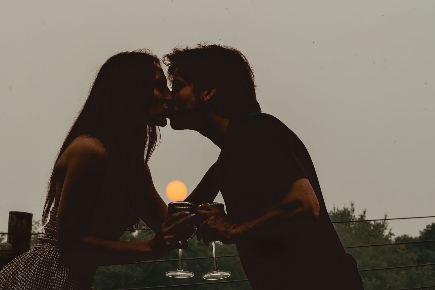 Romantic couple silhouetted against sunset sky sharing a kiss while holding wine glasses outdoors.