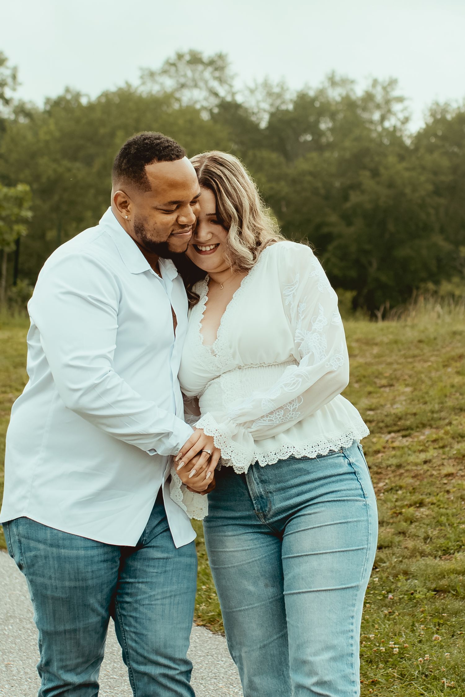 A couple in white tops and jeans share a tender embrace outdoors, smiling and holding hands on a path.