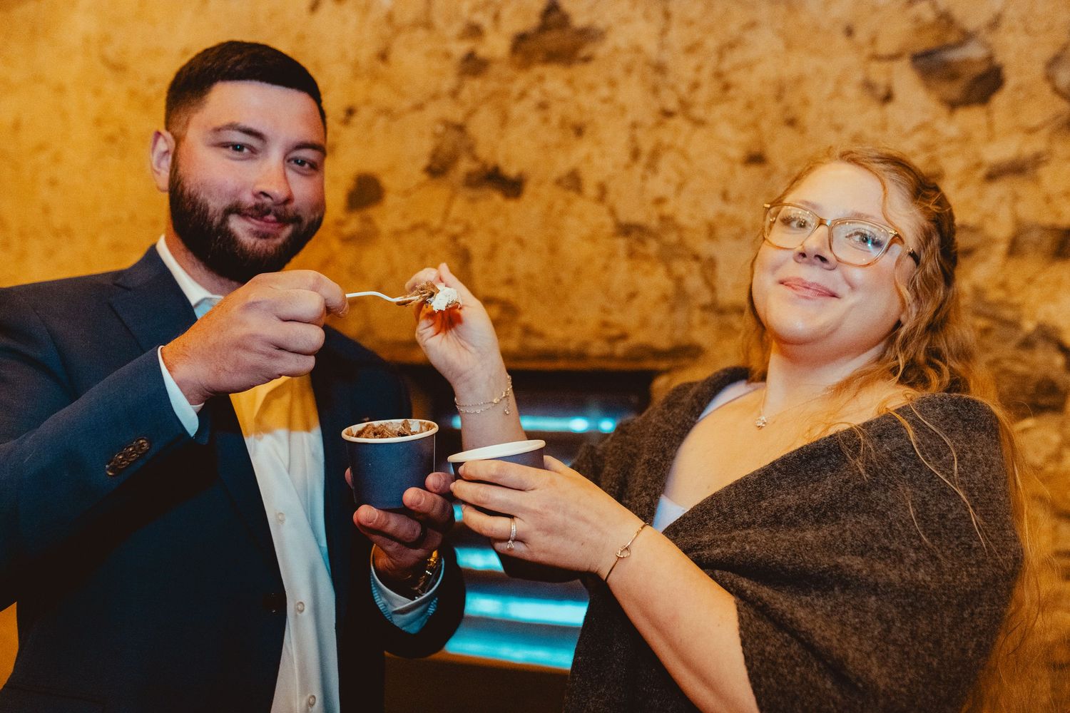 Smiling couple sharing a dessert cup at a rustic stone-walled wedding reception venue.
