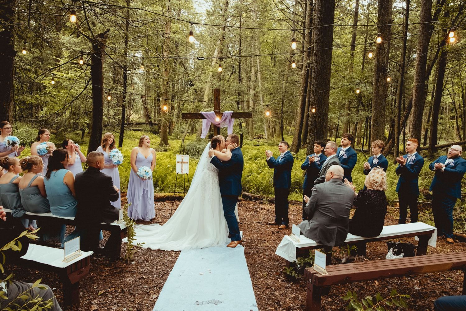Bride and groom share first kiss at outdoor forest wedding ceremony with string lights and wooden benches.
