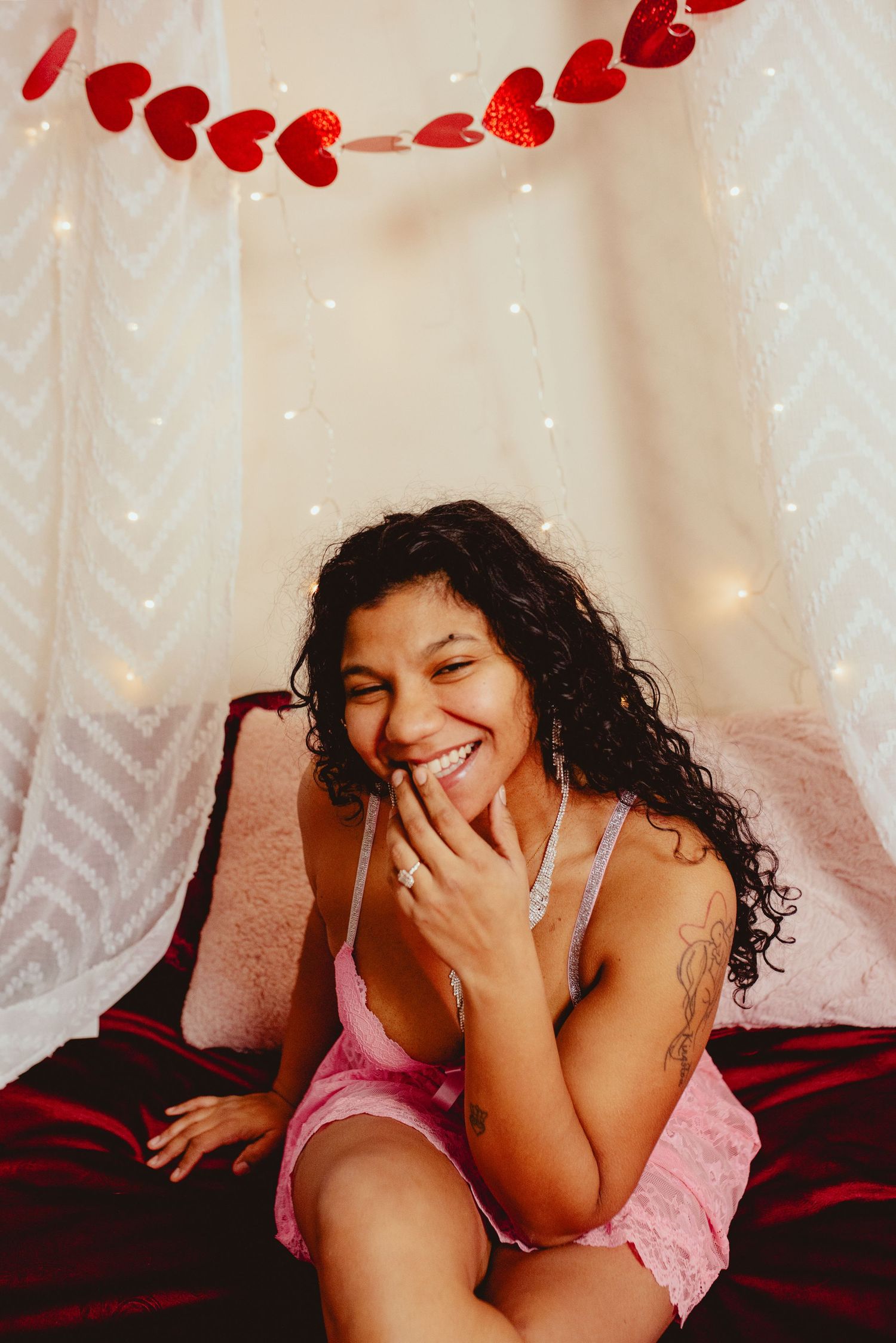 A smiling woman with curly hair sits on a bed decorated with red heart garland and fairy lights.