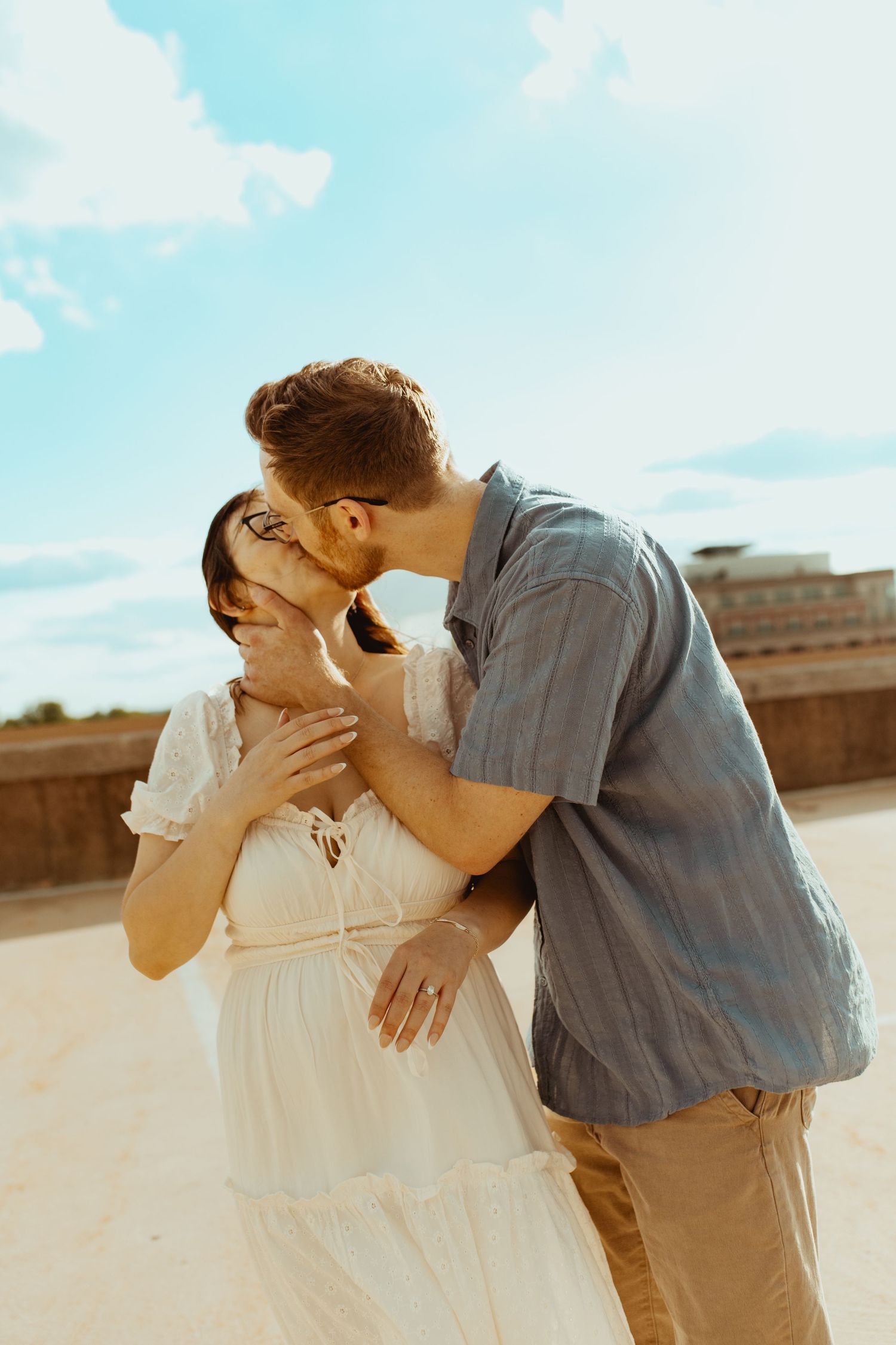 Couple sharing a tender kiss outdoors, woman in white dress and man in blue shirt under a blue sky.