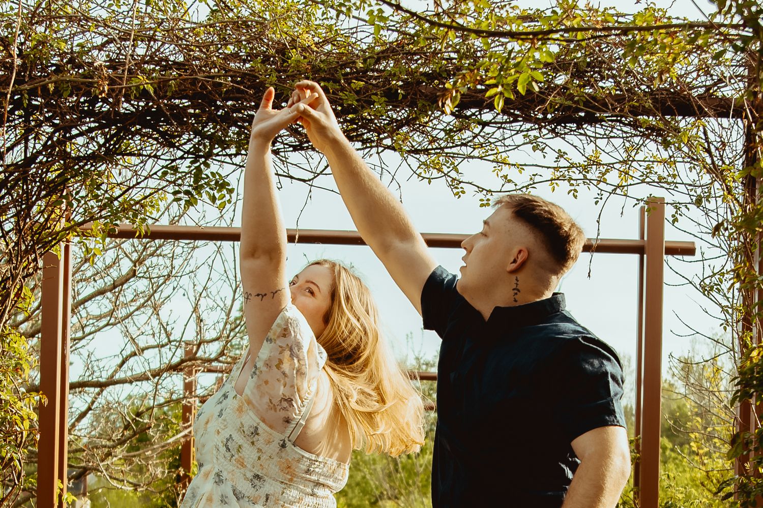 A couple dances under a wooden arbor covered in green vines on a sunny day outdoors.