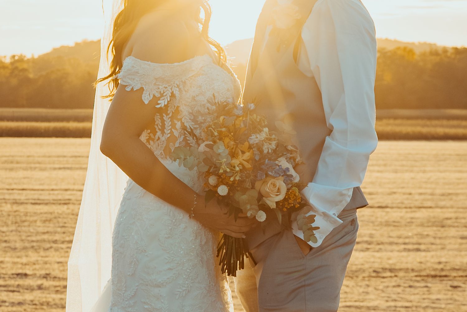 Couple in wedding attire holds hands during golden hour sunset in a field with soft warm lighting.
