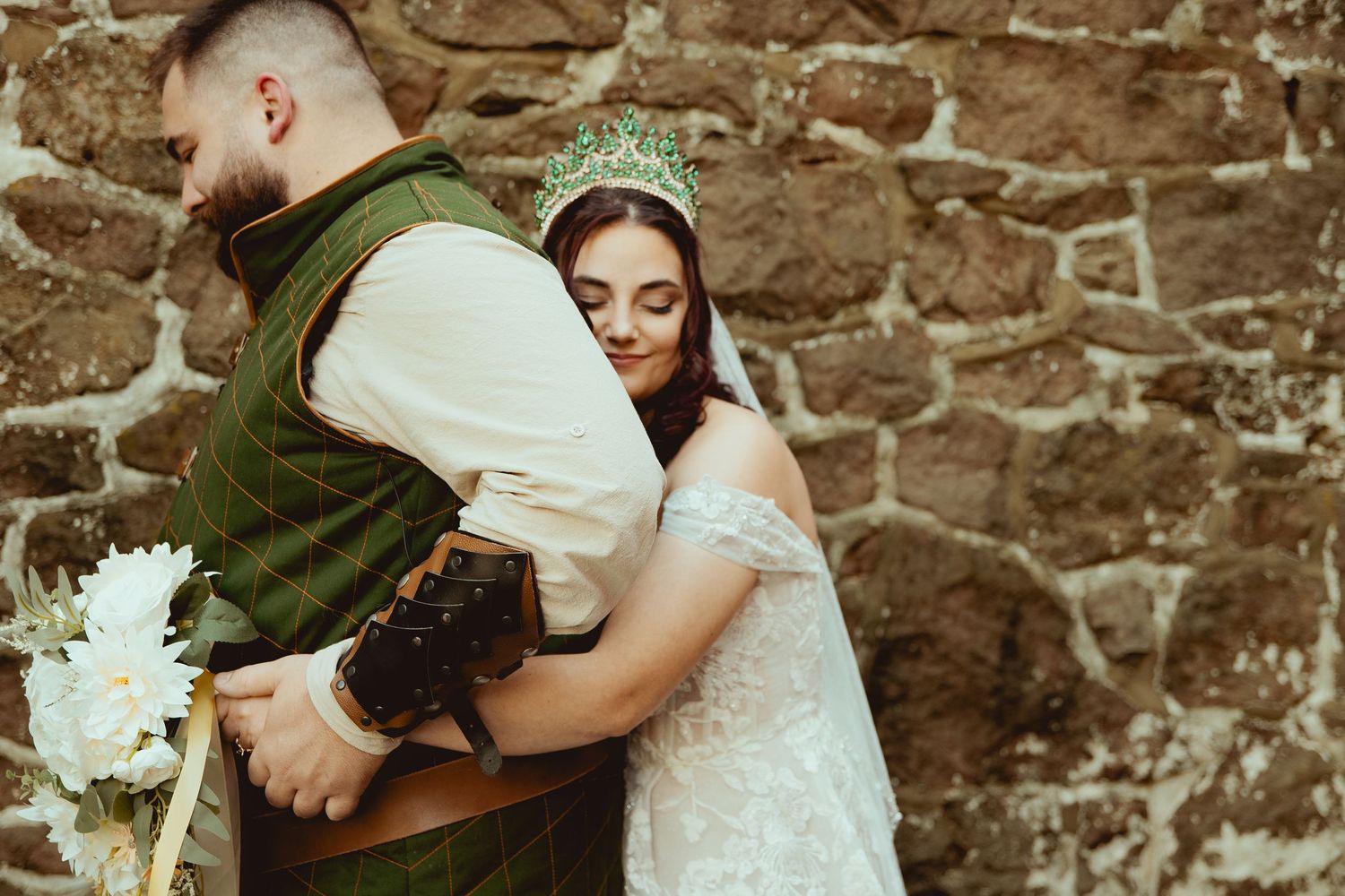 A couple in wedding attire embrace against a rustic stone wall, with the bride wearing a tiara and lace dress.