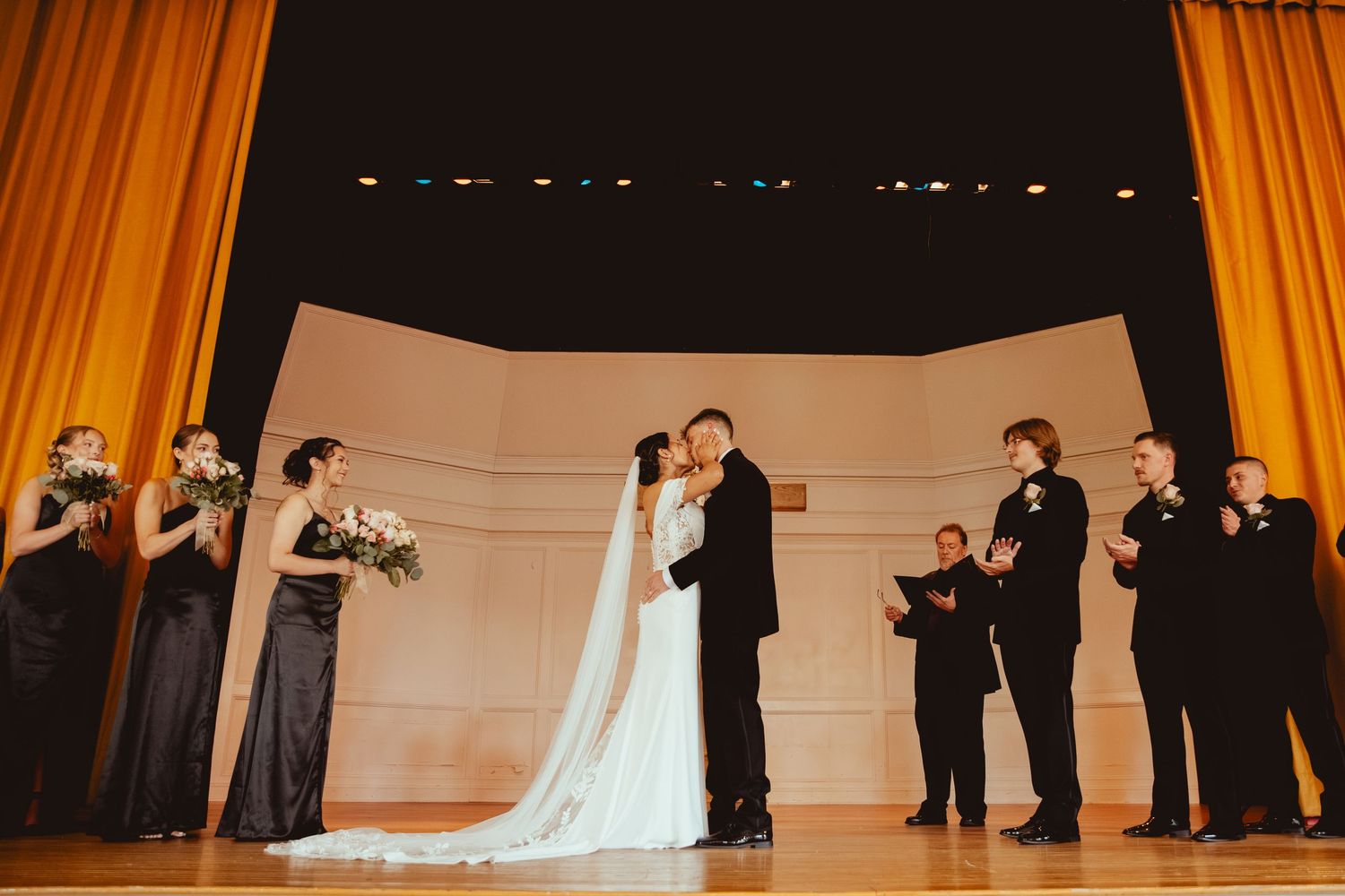 Wedding ceremony in progress on stage with orange curtains and floral arrangements.