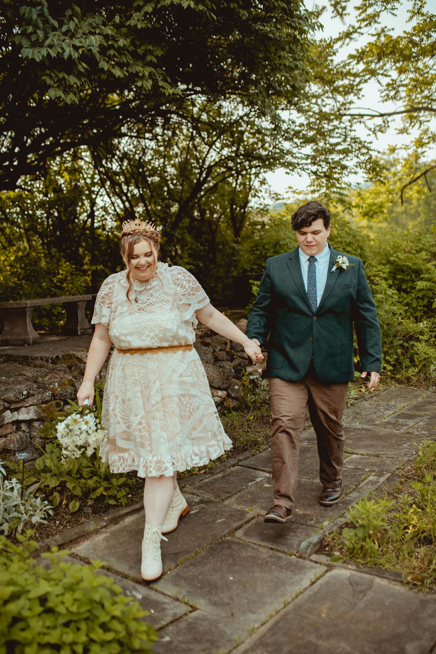 A couple walks hand in hand along a rustic stone path surrounded by green foliage during their wedding.