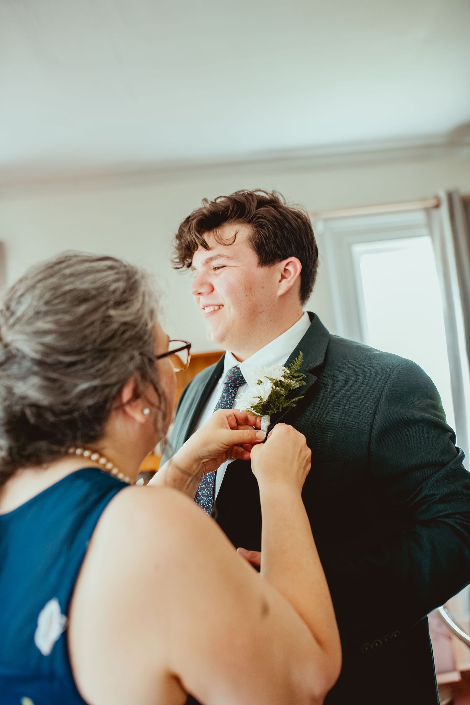A person in a navy suit and boutonniere smiles while someone in a blue dress adjusts their floral lapel pin.