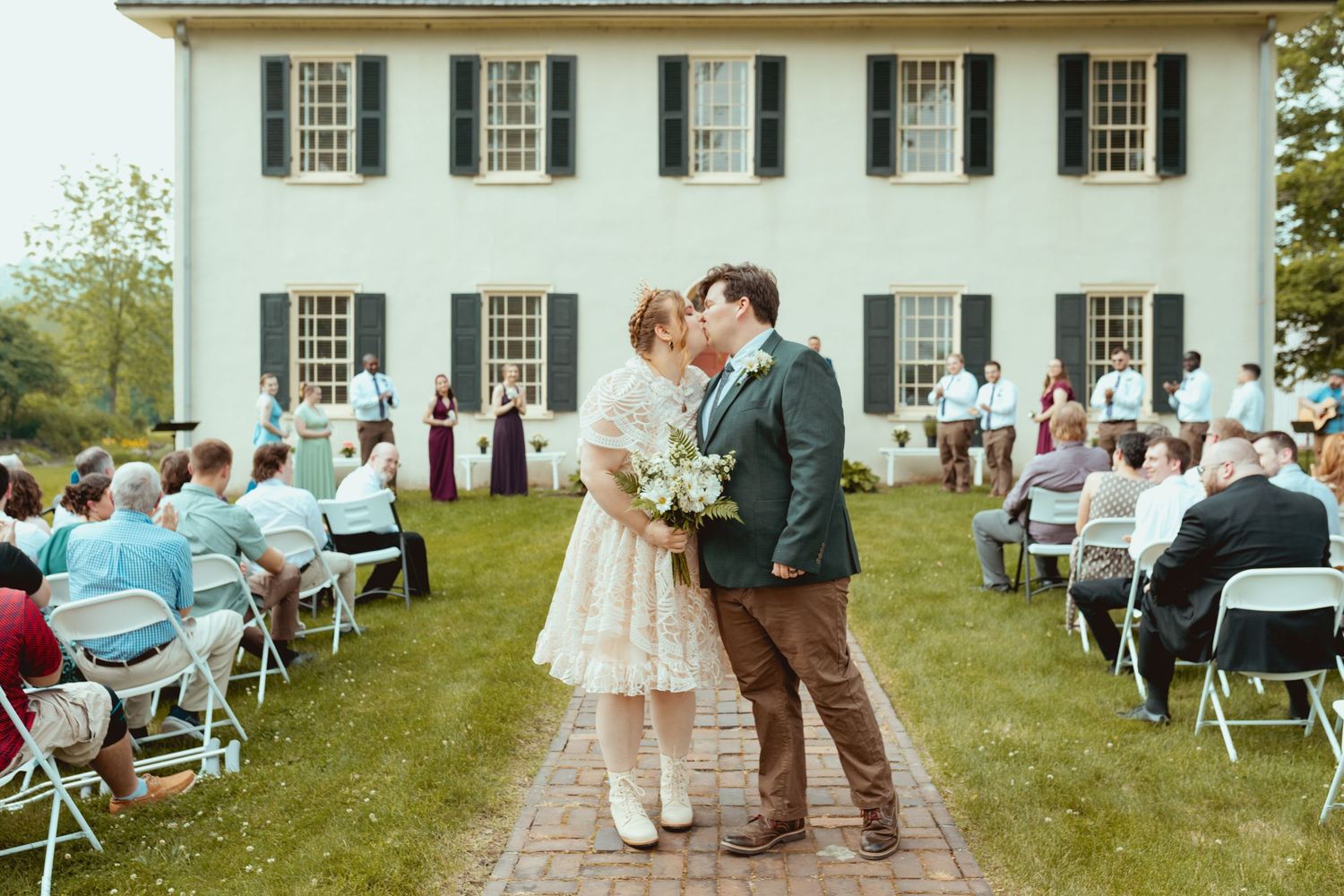 A couple shares a romantic moment on a brick path during an outdoor wedding ceremony with white chairs.