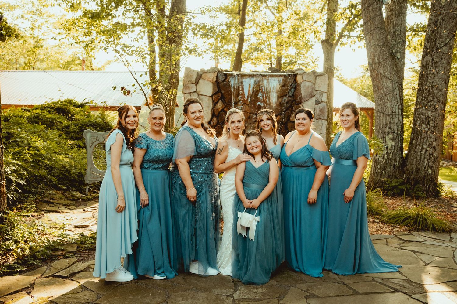 Bridal party in coordinating teal dresses and suits poses outdoors against a sunlit wooded backdrop.