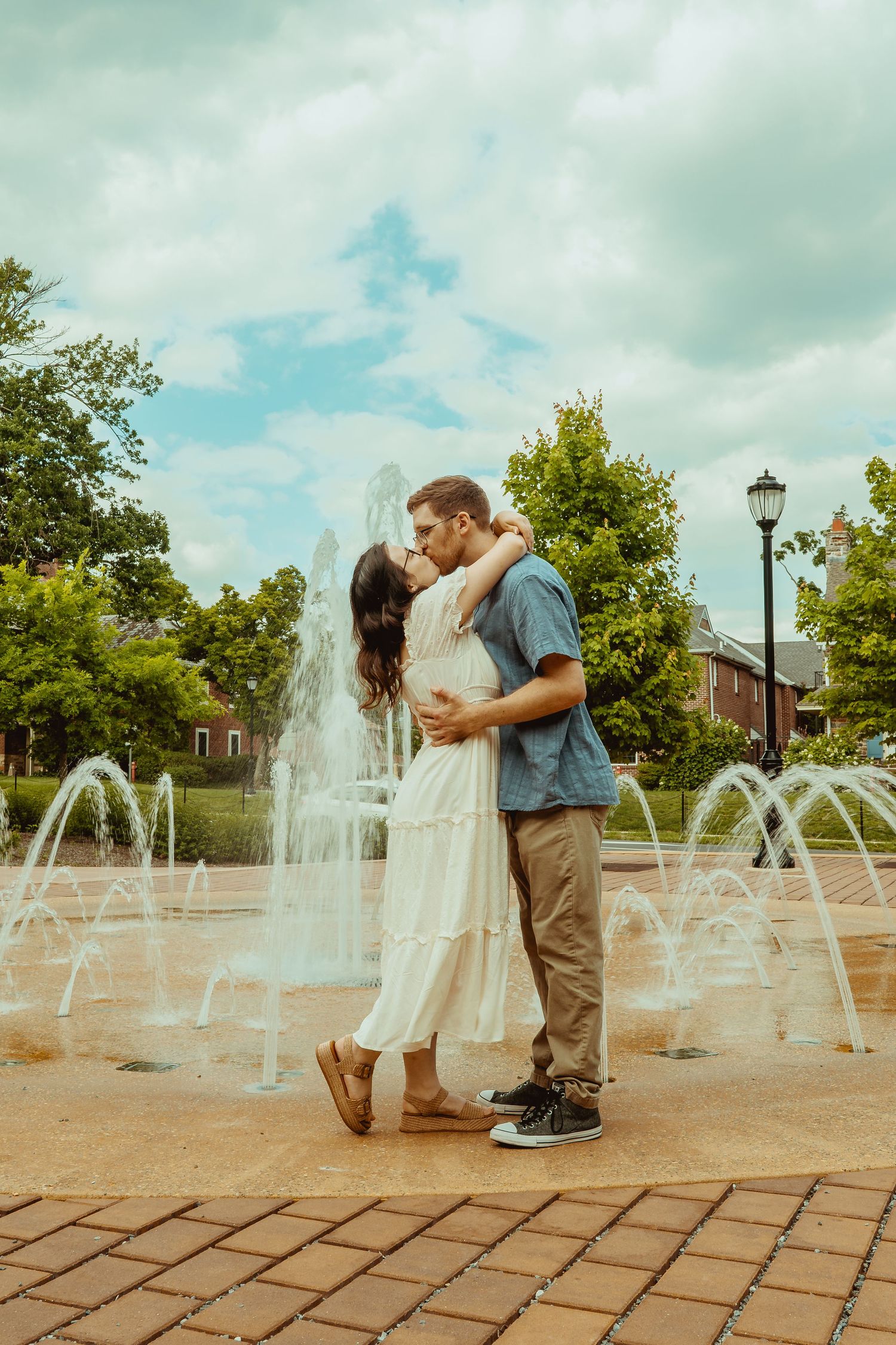 Couple sharing a romantic kiss in front of a fountain in a park, woman in white dress, man in blue shirt.