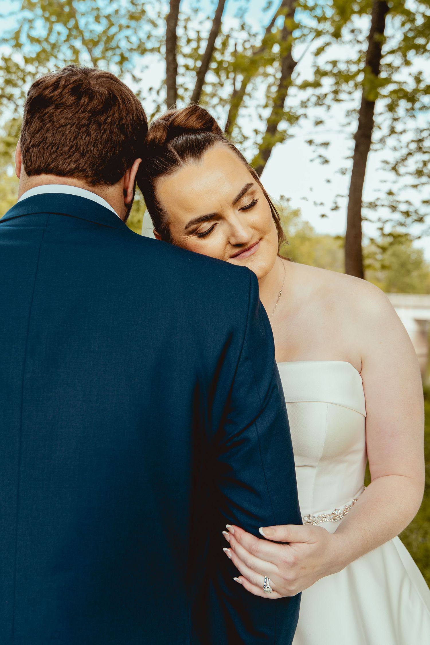 Bride in strapless white gown resting head on grooms navy suit shoulder in romantic outdoor moment.
