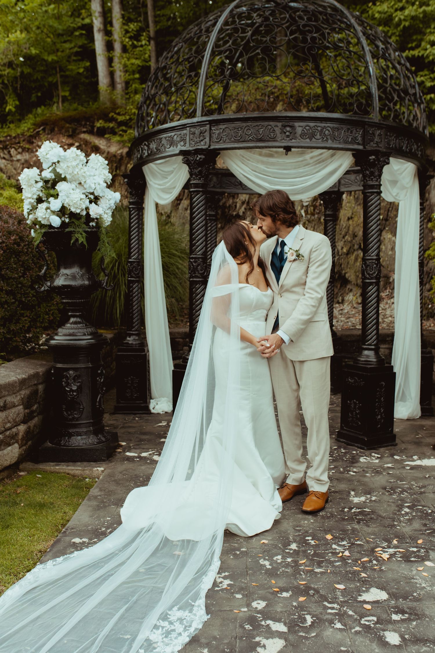 Bride and groom share first kiss under ornate black gazebo with white draping and floral arrangements.