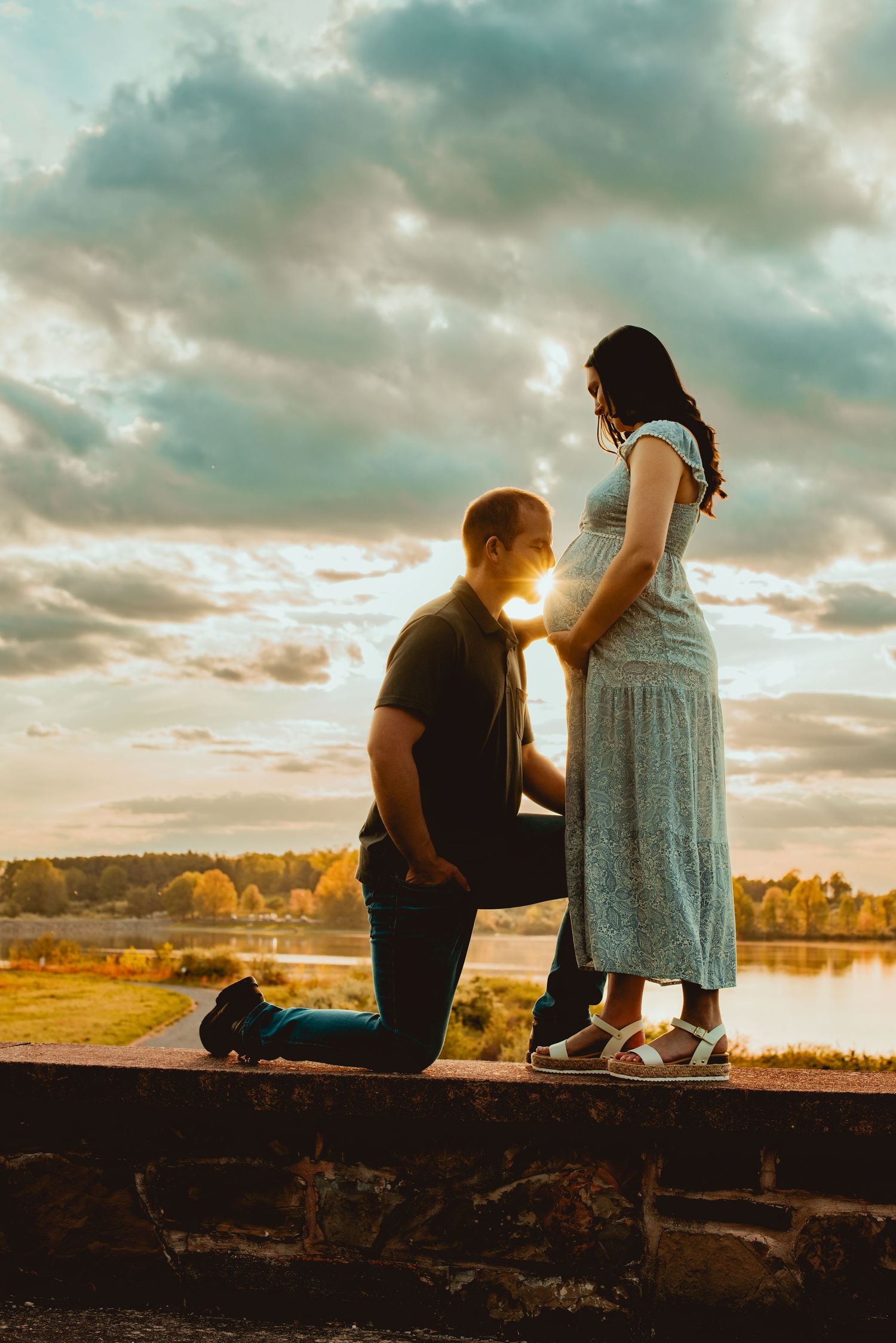 A couple sits together on a wooden ledge at sunset overlooking a serene lake with dramatic clouds above.