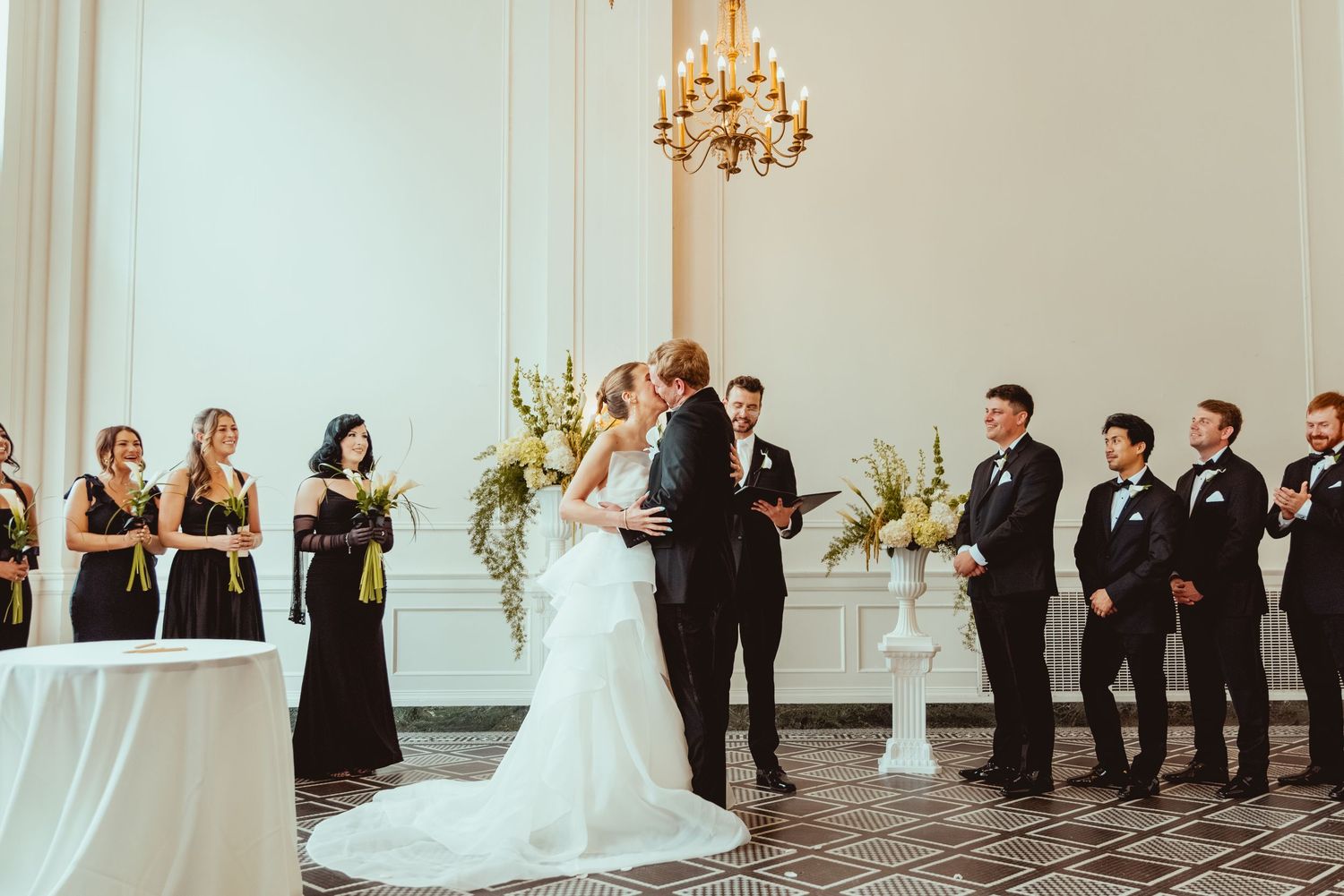 Wedding party stands in formation along white walls of an elegant ballroom during ceremony.