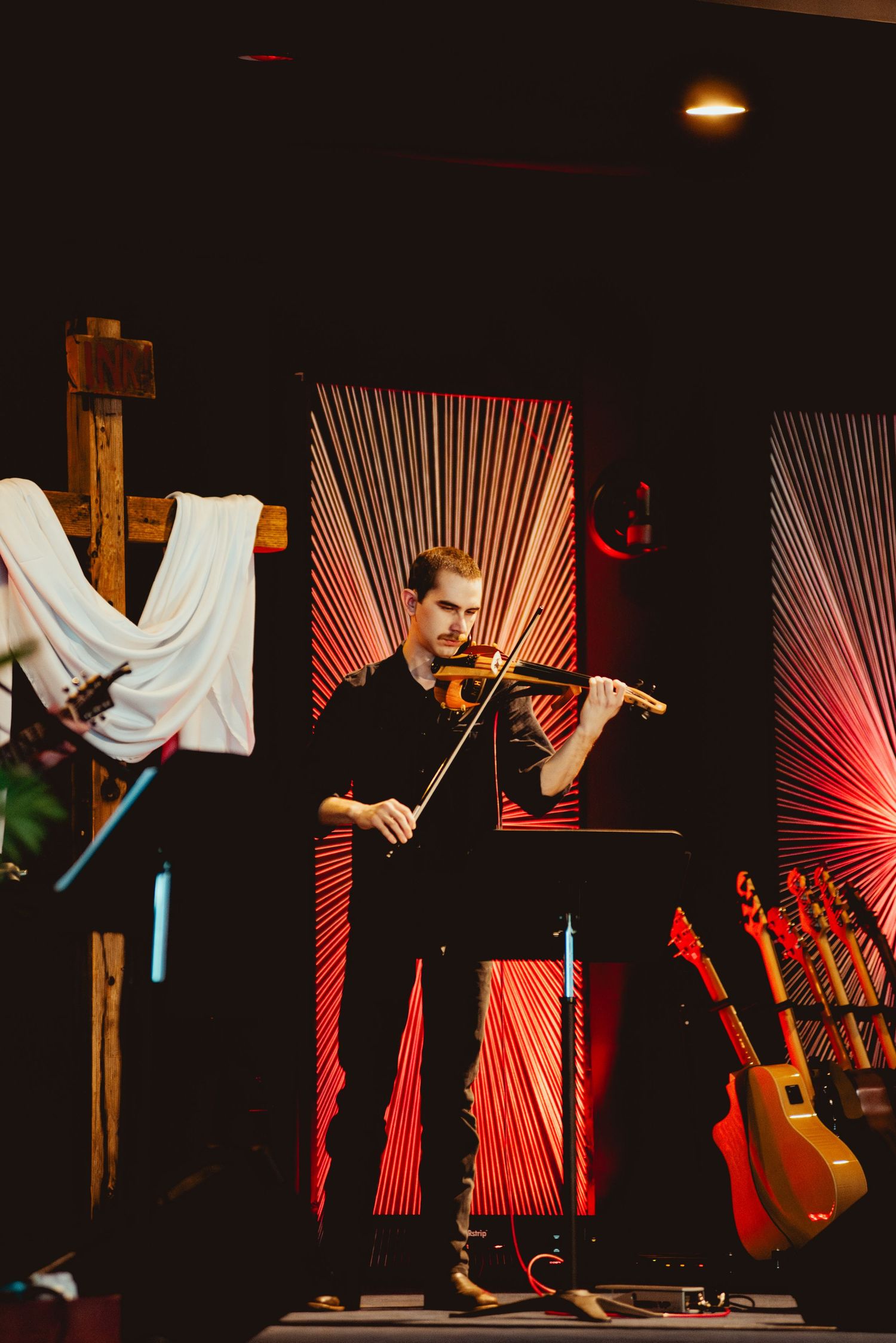 A violinist performs on stage with dramatic red string light patterns illuminating the dark background.