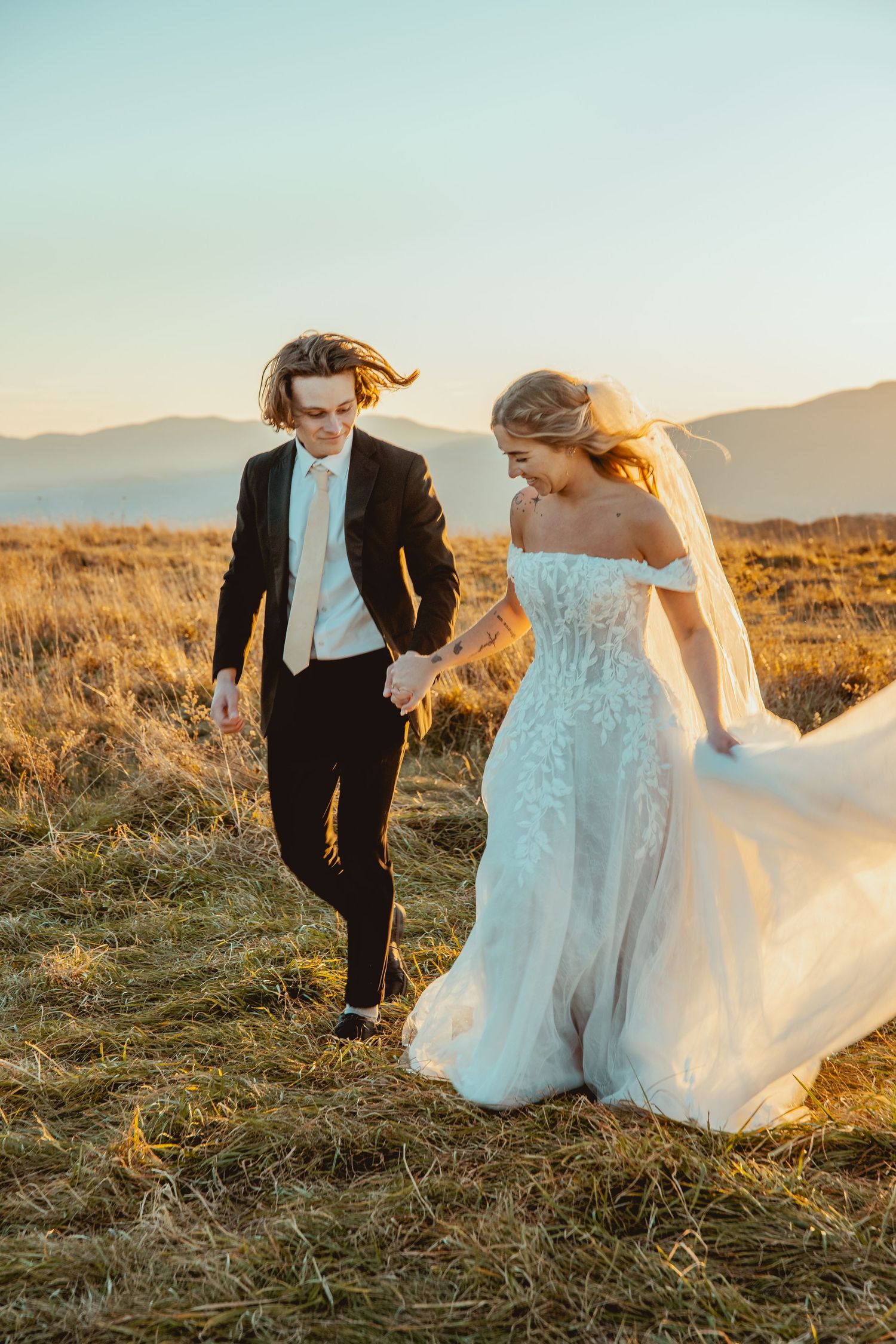 Wedding couple walks hand in hand through golden grass at sunset with mountains in the distance.