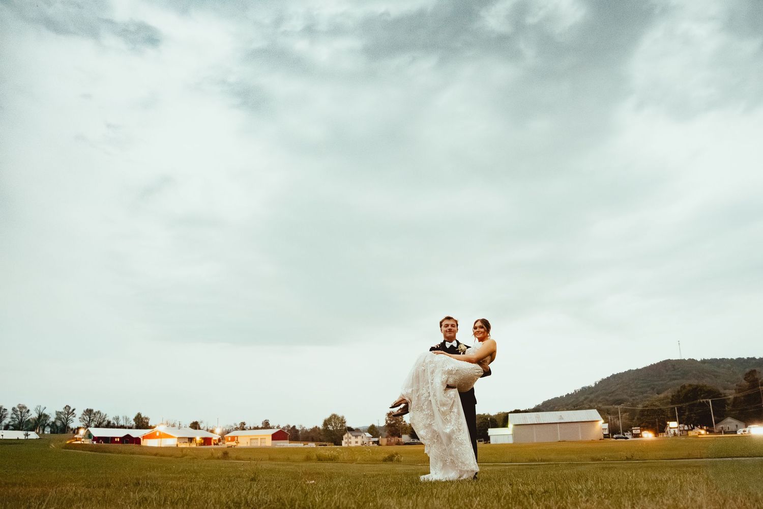 A couple in wedding attire stands in a scenic field under cloudy skies during a romantic photo session.
