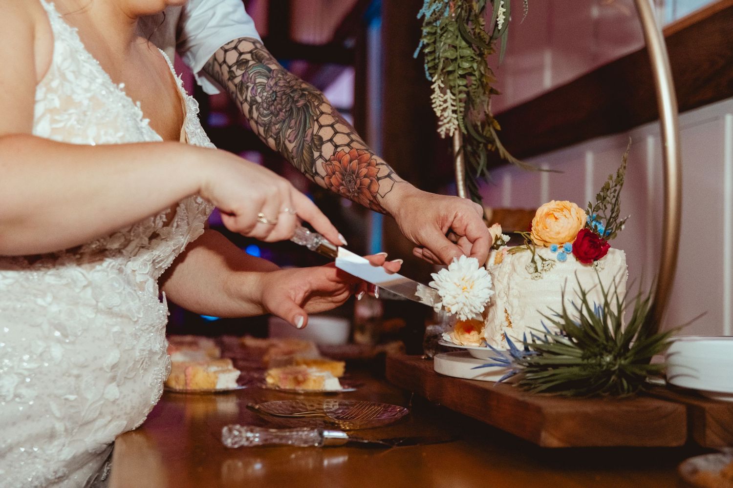 Couple cutting a floral wedding cake together, one partner wearing a lace gown and the other showing tattooed arms.