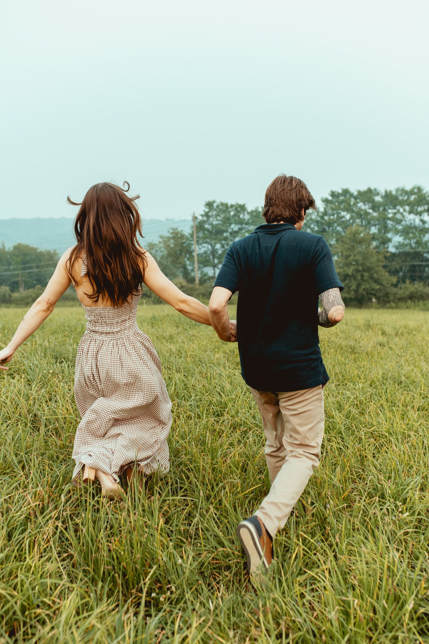 Couple holding hands running through a lush green field, woman in beige dress and man in navy shirt.
