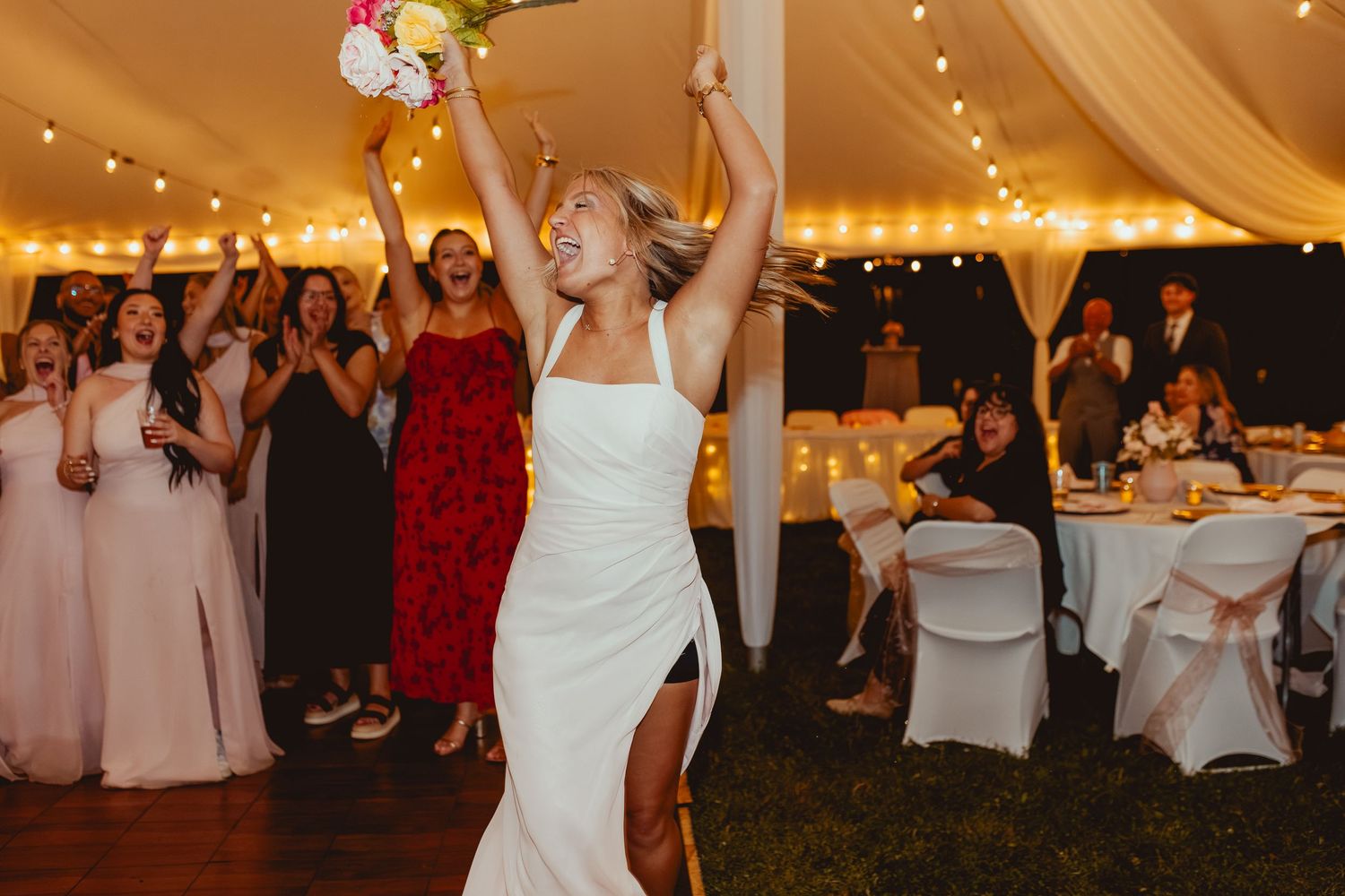 Bride tosses bouquet to excited guests under string lights at outdoor wedding reception.