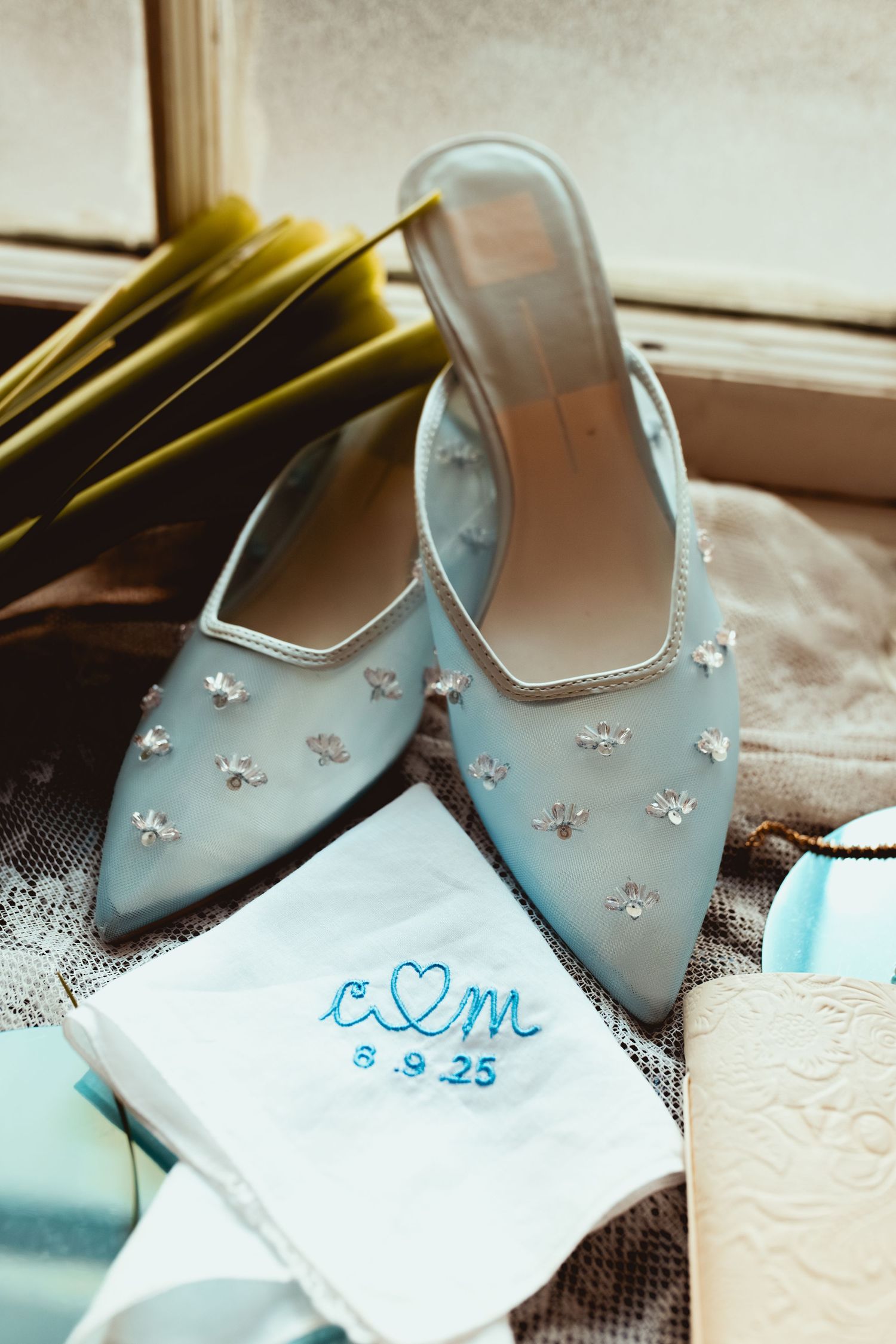 Light blue embellished bridal mules with silver beading displayed next to a monogrammed white handkerchief.