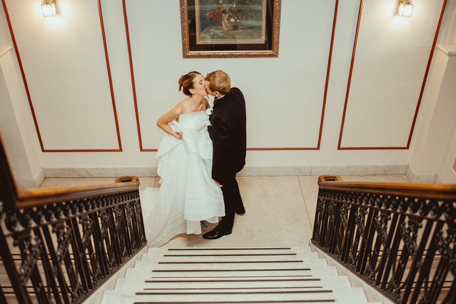 A couple in wedding attire share a romantic moment at the top of an elegant staircase with ornate railings.