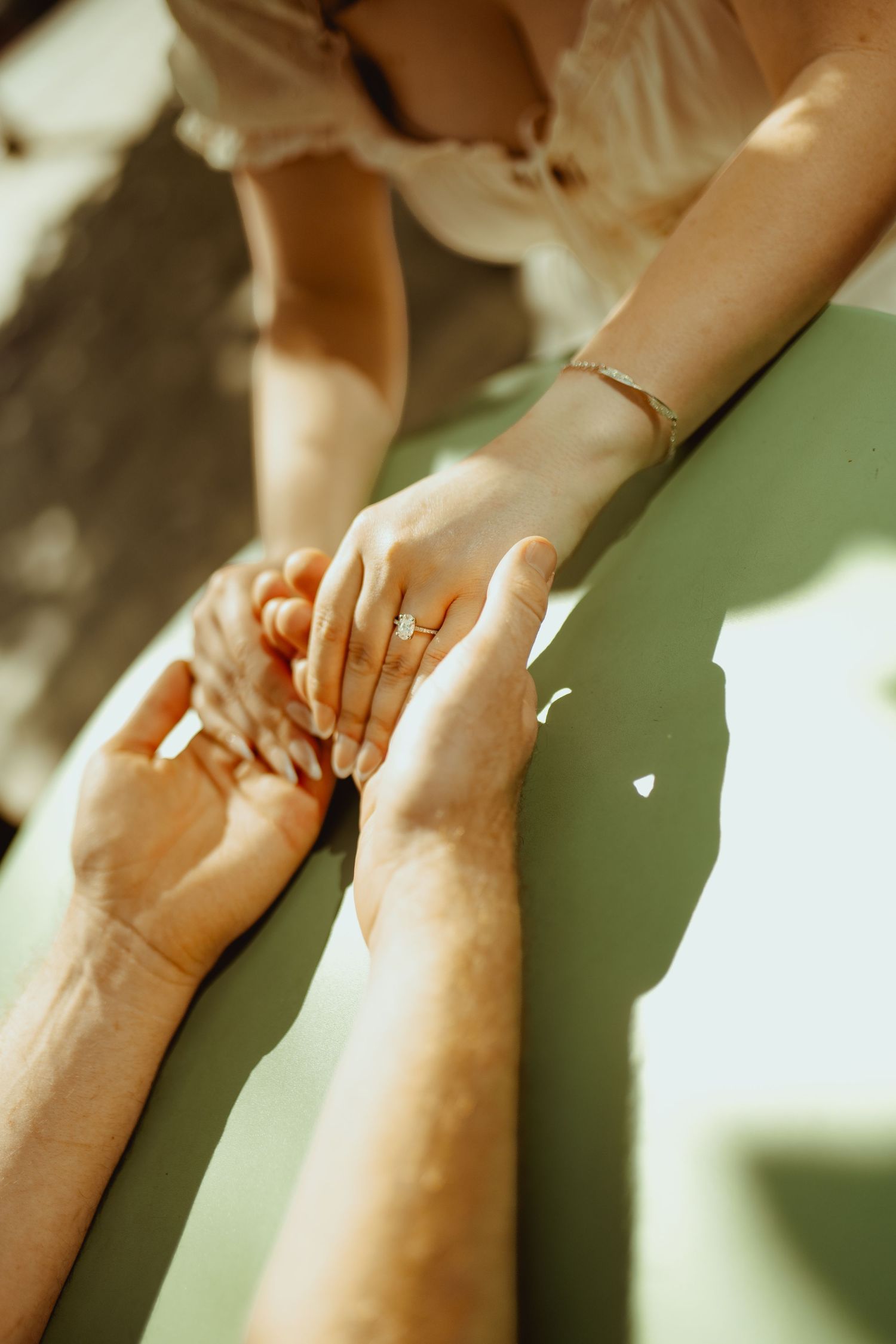 Close-up of a couple holding hands, woman wearing a diamond engagement ring and gold bracelet in warm sunlight.