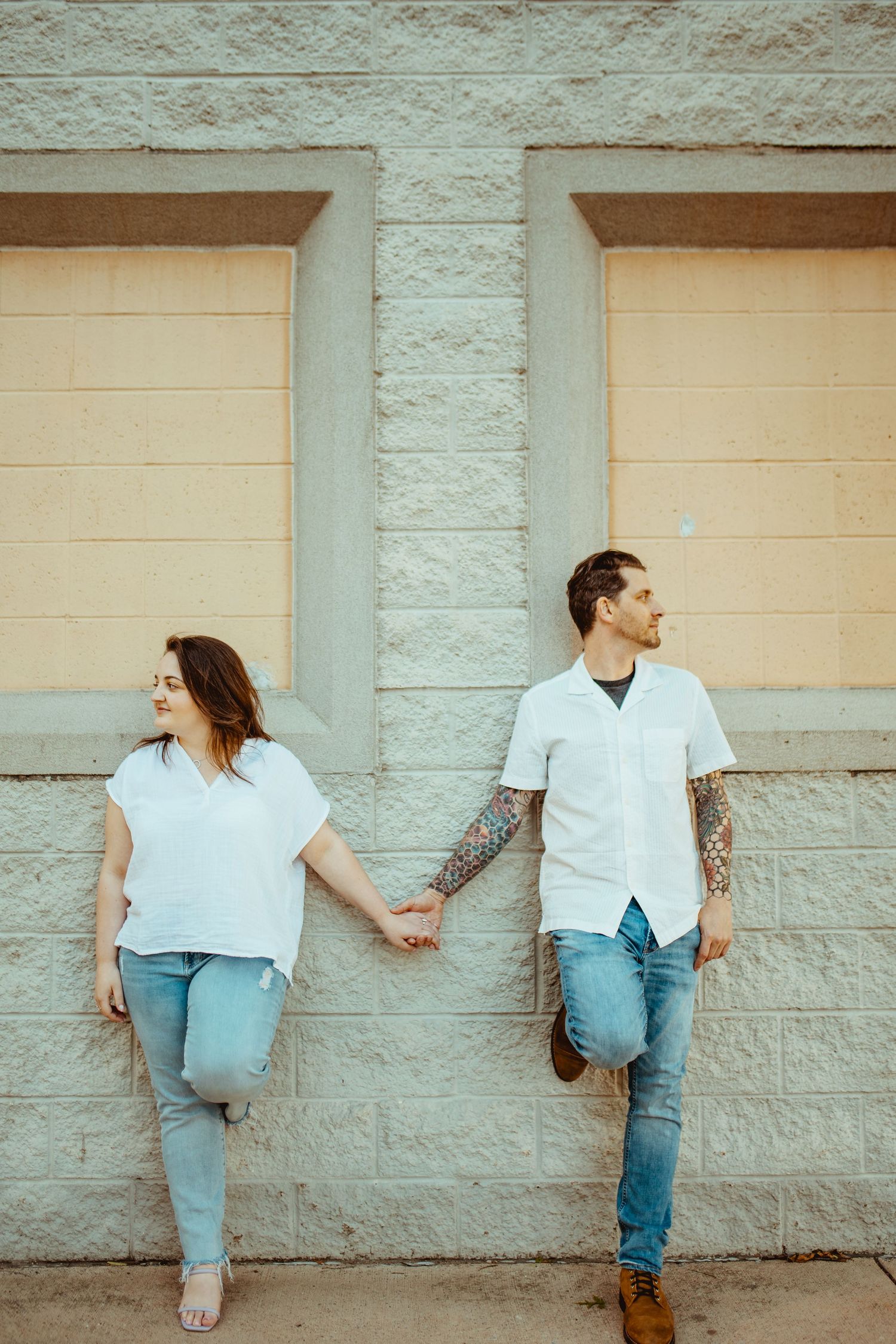 Couple holding hands leaning against stone wall in matching white shirts and jeans for engagement photos.